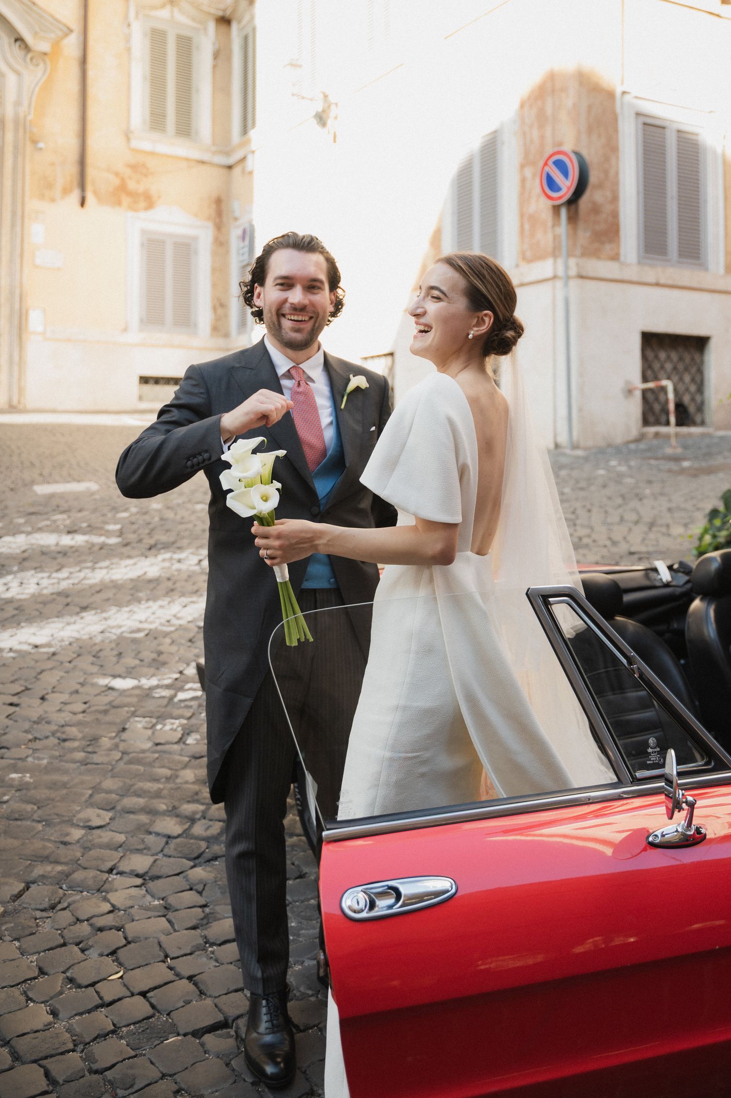 A joyful wedding couple poses next to a red convertible on Italian cobblestone streets.