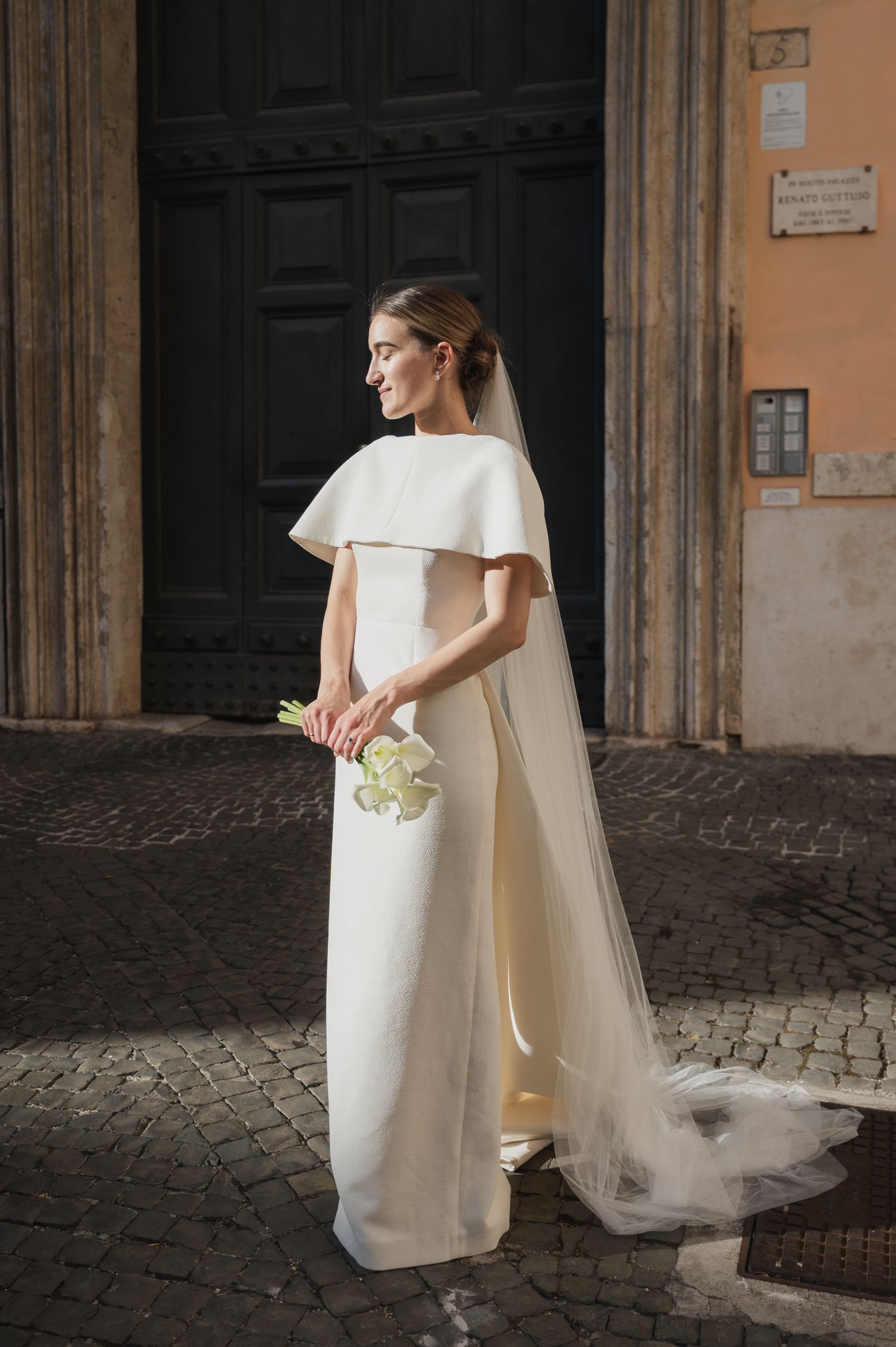 Modern bride with calla lily bouquet standing against historic European building facade.
