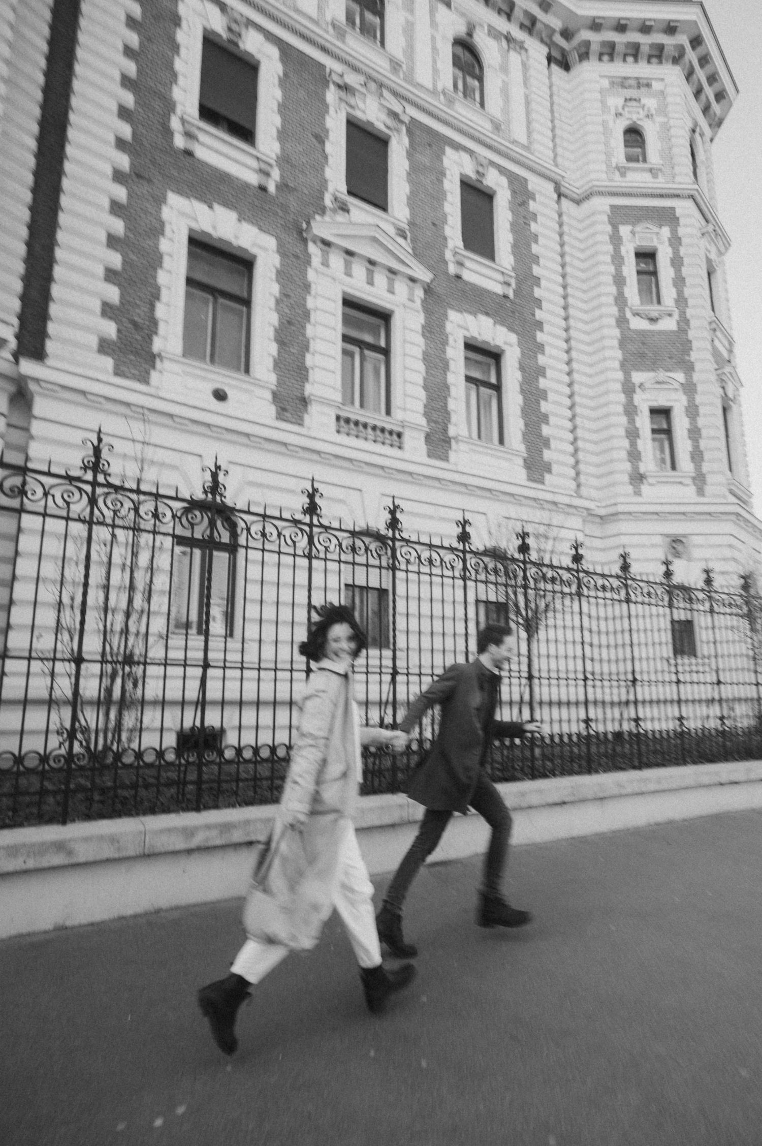 Two people walking past an ornate historic building with wrought iron fencing in black and white photography.