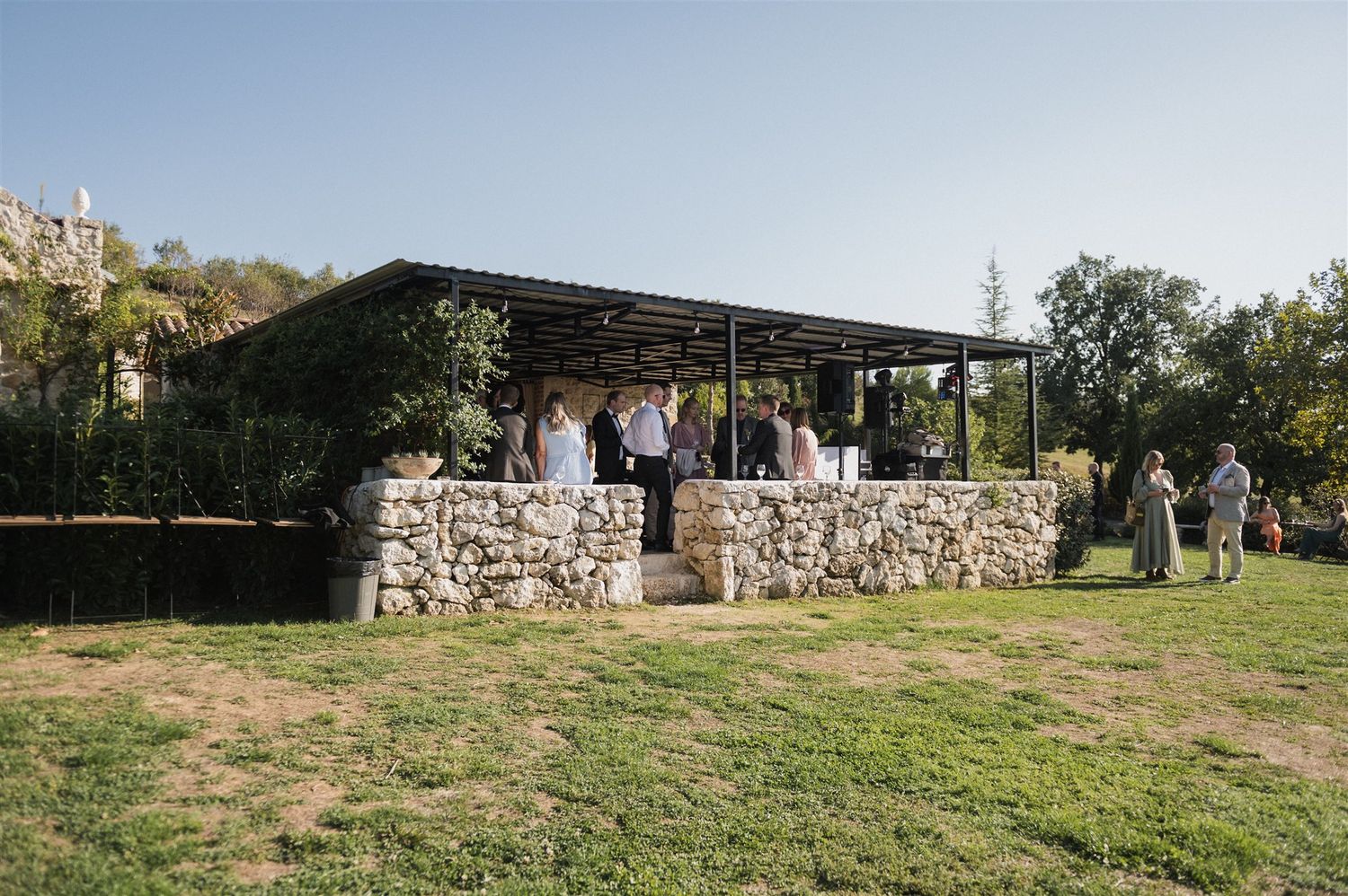 Wedding guests mingle under a stone pavilion structure in an outdoor garden setting.