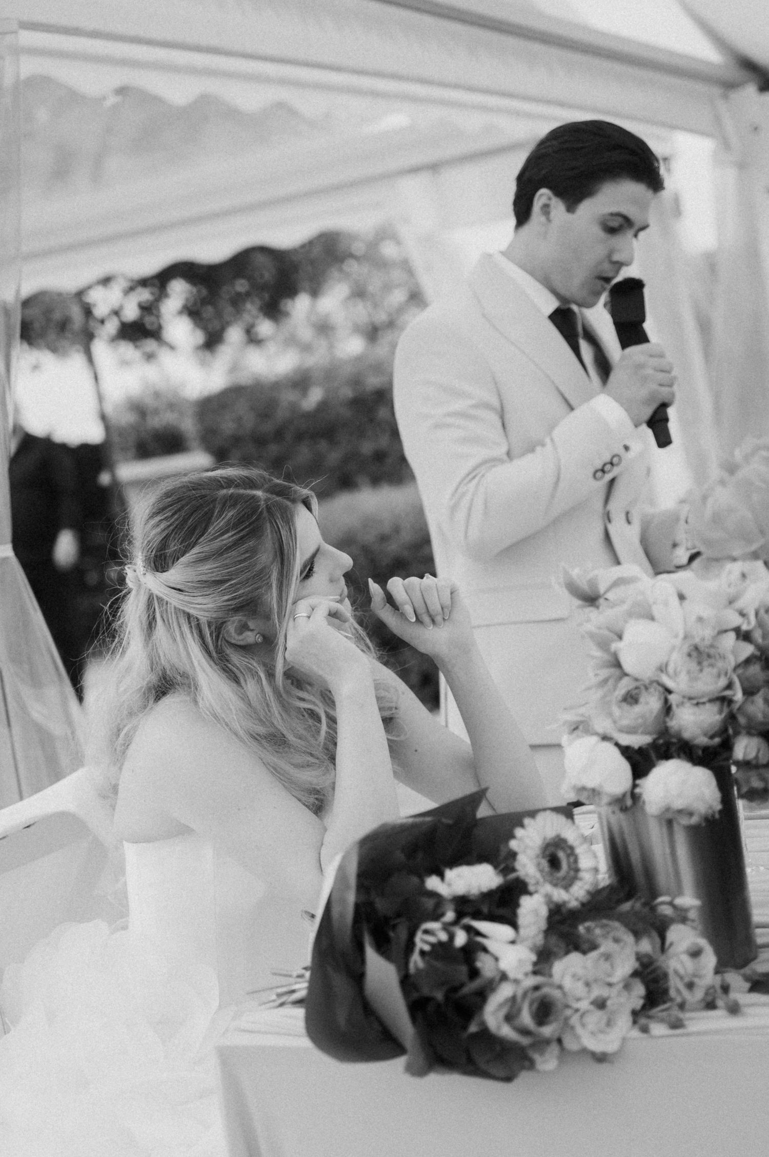 Emotional bride wipes tears as groom in white suit delivers heartfelt wedding speech, black and white.