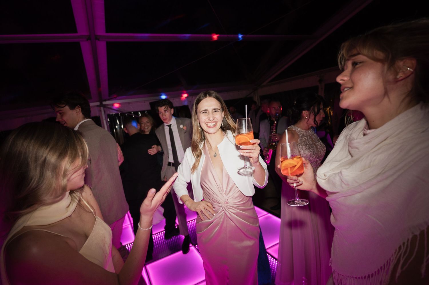 Woman in pink dress smiles and holds a cocktail while dancing at an evening wedding reception party.