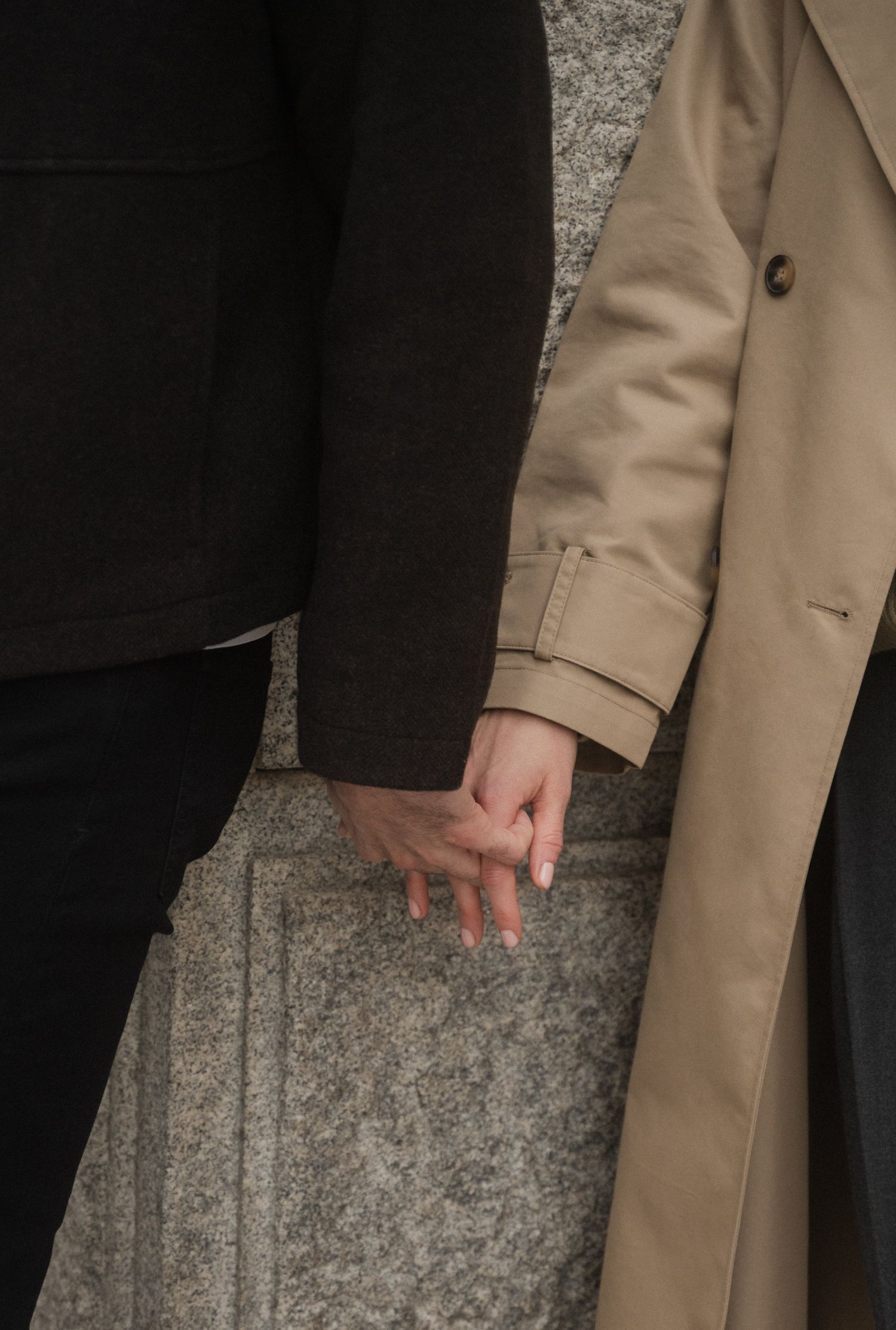 A person wearing a beige trench coat and black pants stands against a textured grey wall.