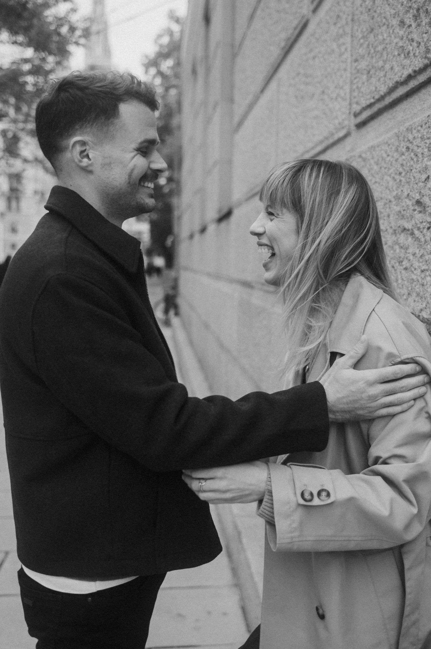 Black and white candid photo of a couple laughing and embracing on a city street beside a textured wall.