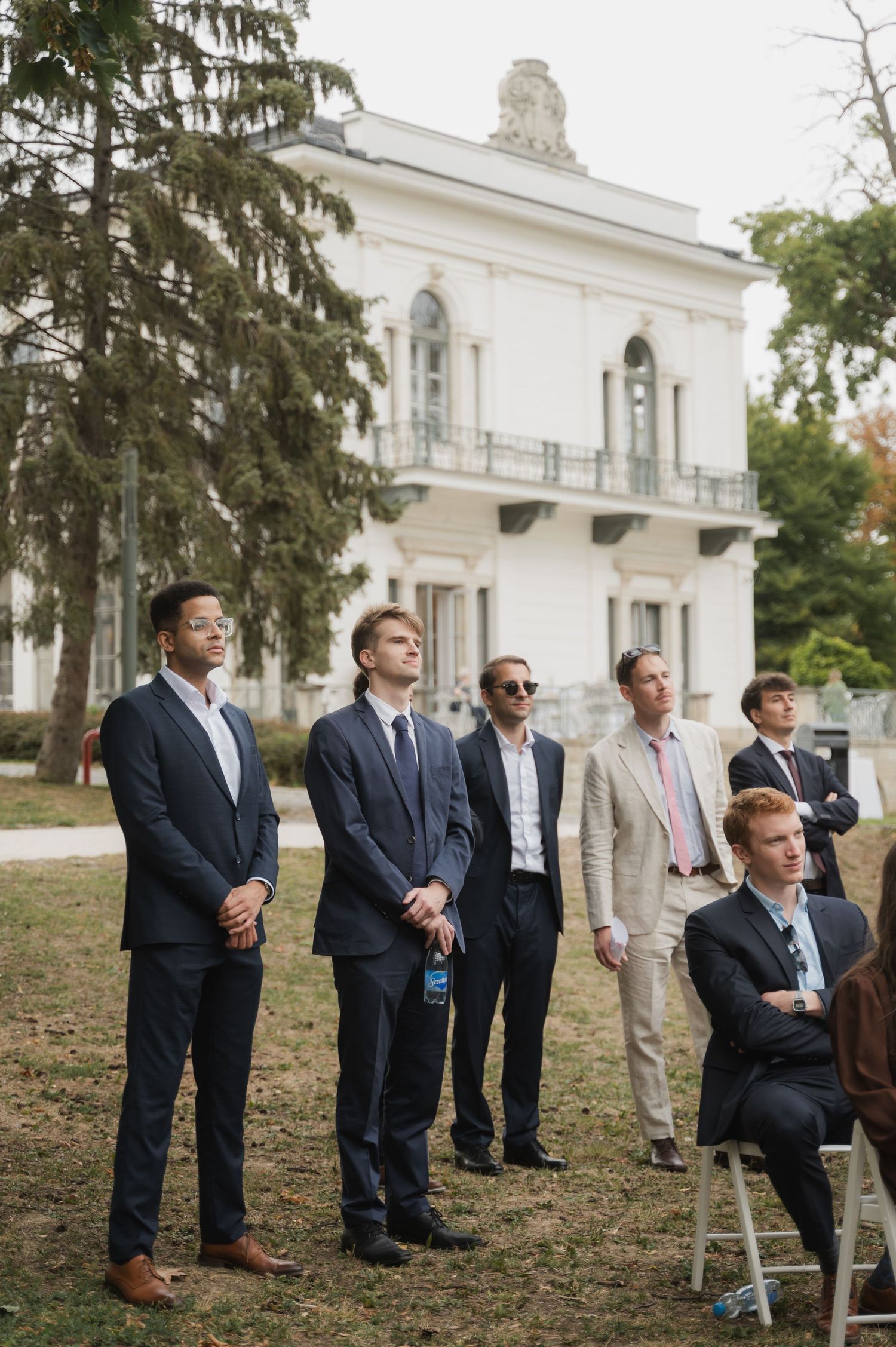 Groomsmen and wedding guests in suits watching outdoor ceremony near white historic mansion.