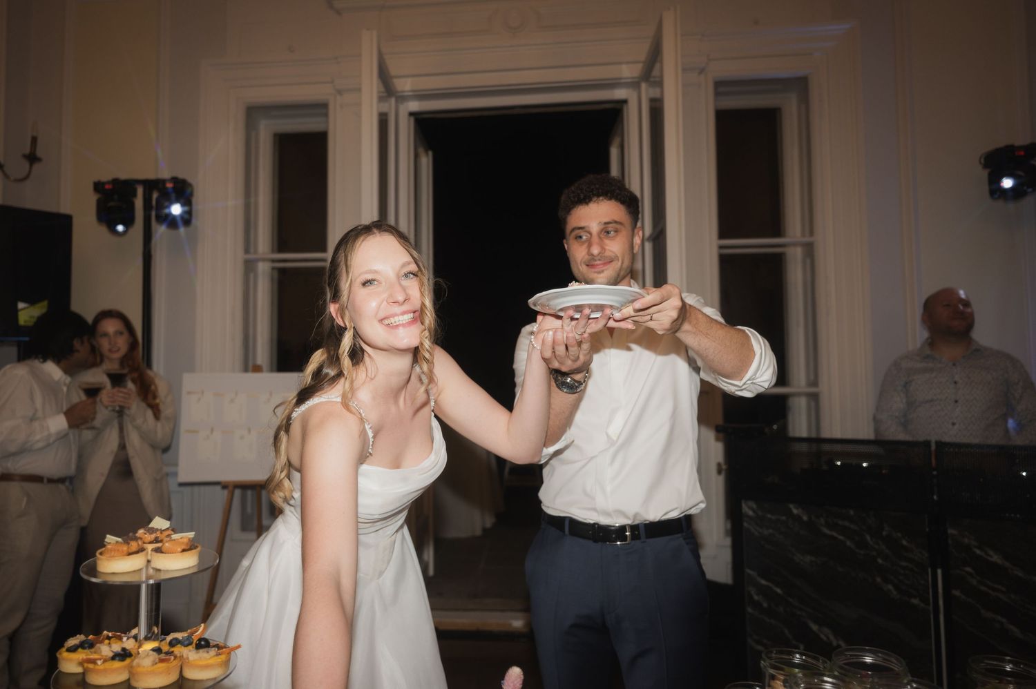 Bride and groom laughing while cutting their wedding cake at an elegant evening reception.