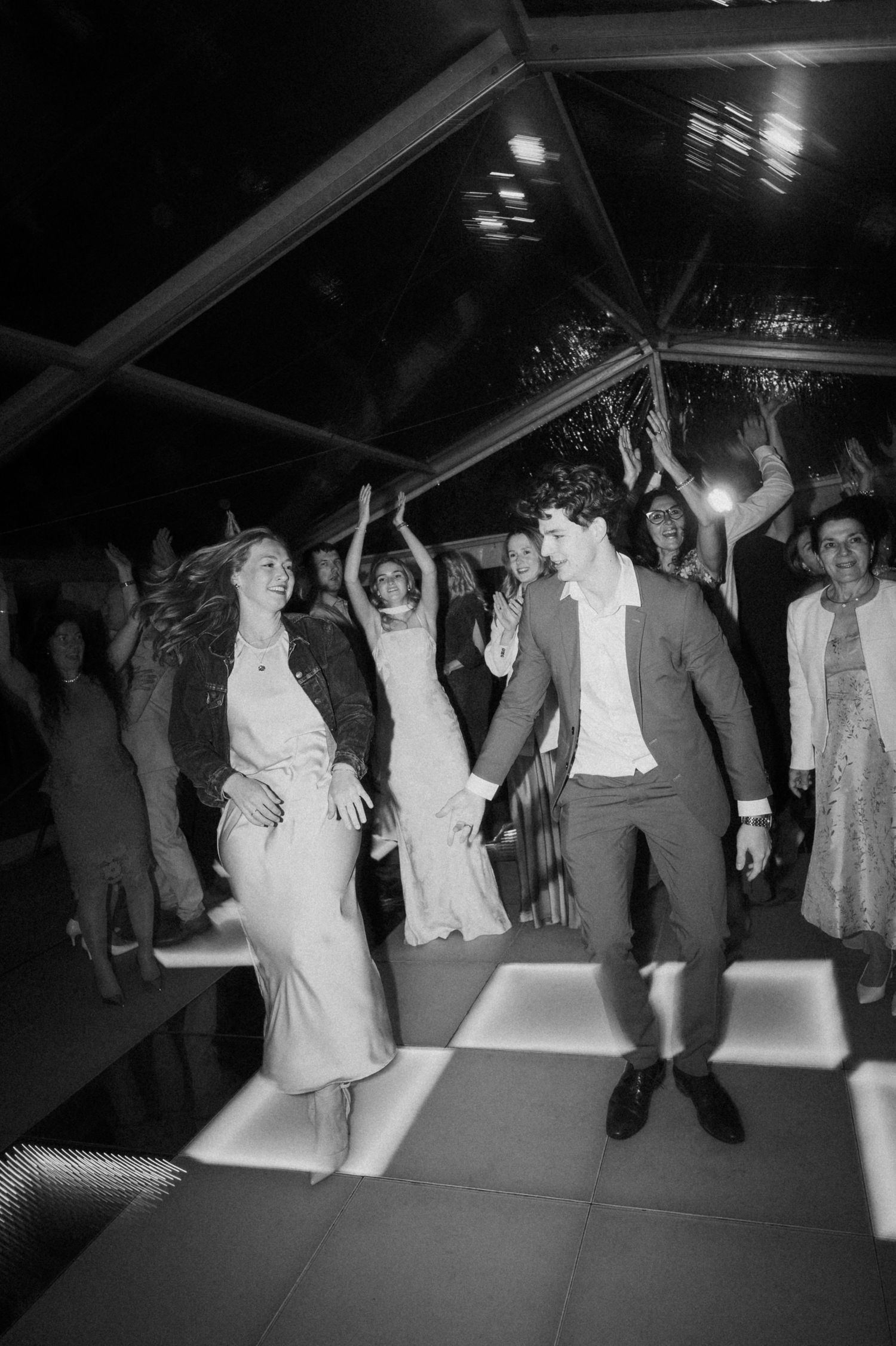 Black and white photo of bride and groom dancing joyfully with guests at a lively wedding reception.