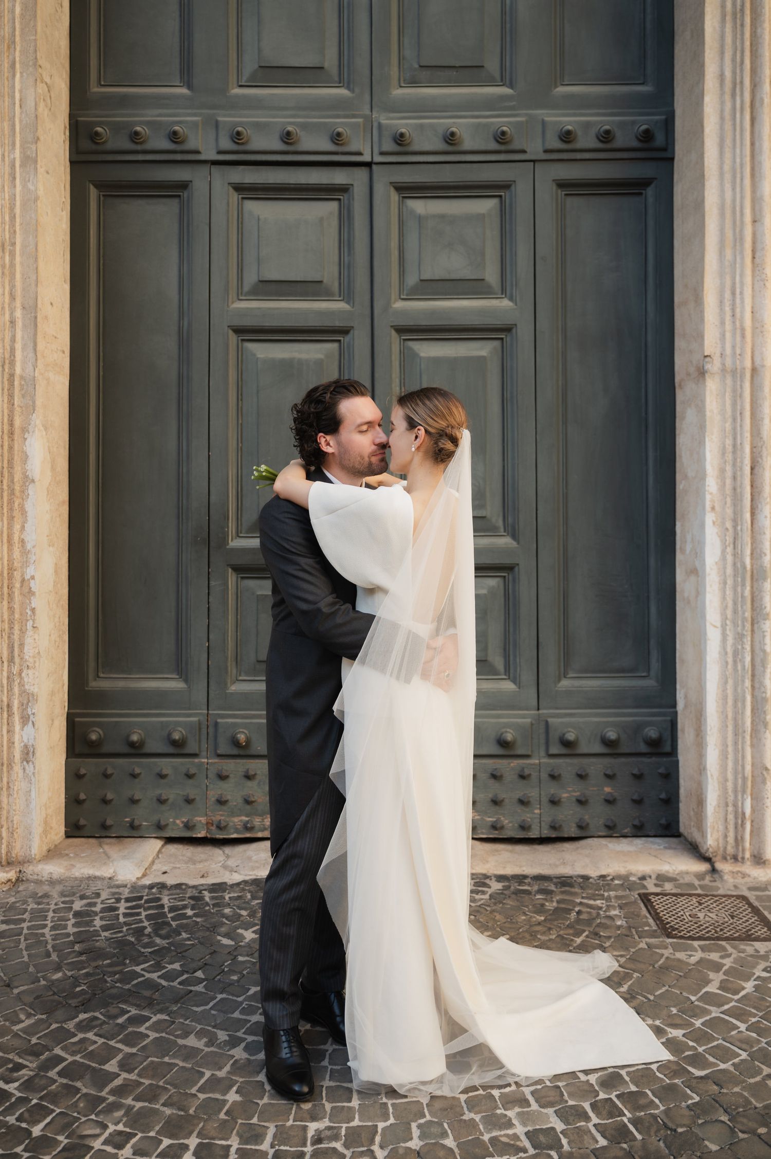 Wedding couple shares intimate moment in front of ornate wooden church doors.