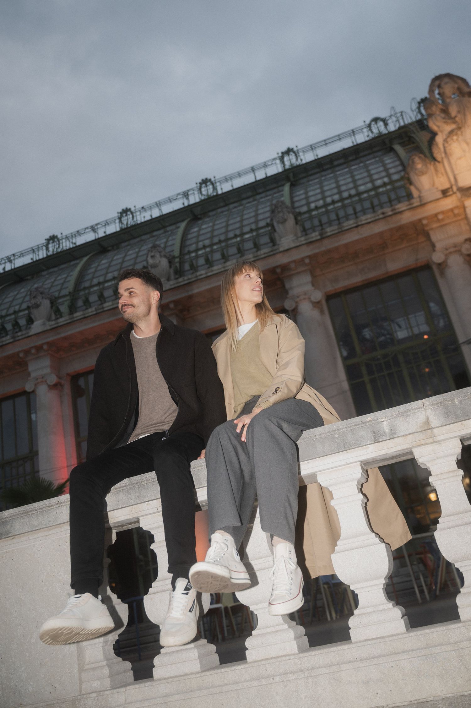 Two people sit casually on ornate stone balustrade against backdrop of classical architecture at dusk.