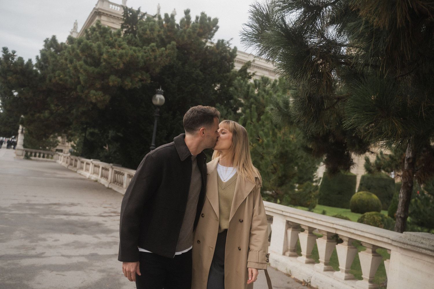 A couple shares a tender moment on a stone balustrade walkway surrounded by palm trees and greenery.