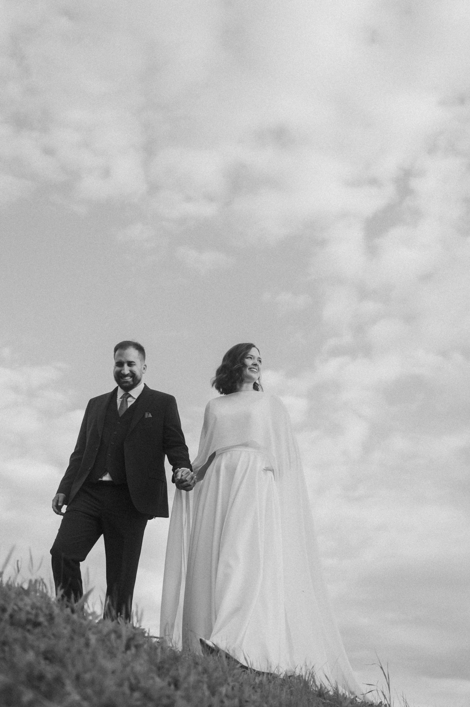 A bride and groom stand together on a hilltop silhouetted against a cloudy sky in a dramatic black and white photo.