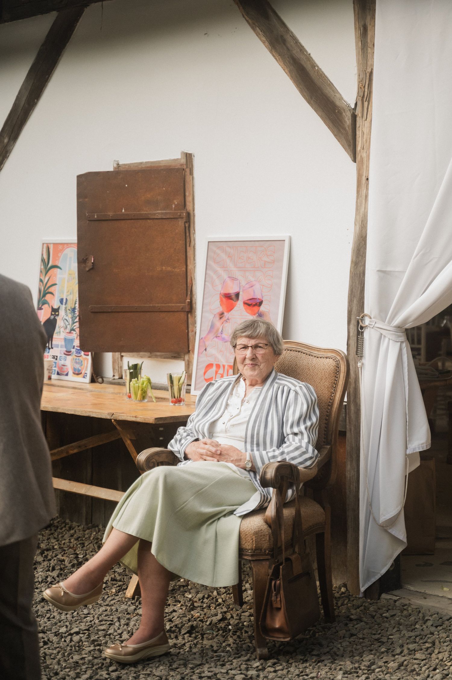 A person sits in a vintage armchair in a rustic room with wooden beams and white curtains.