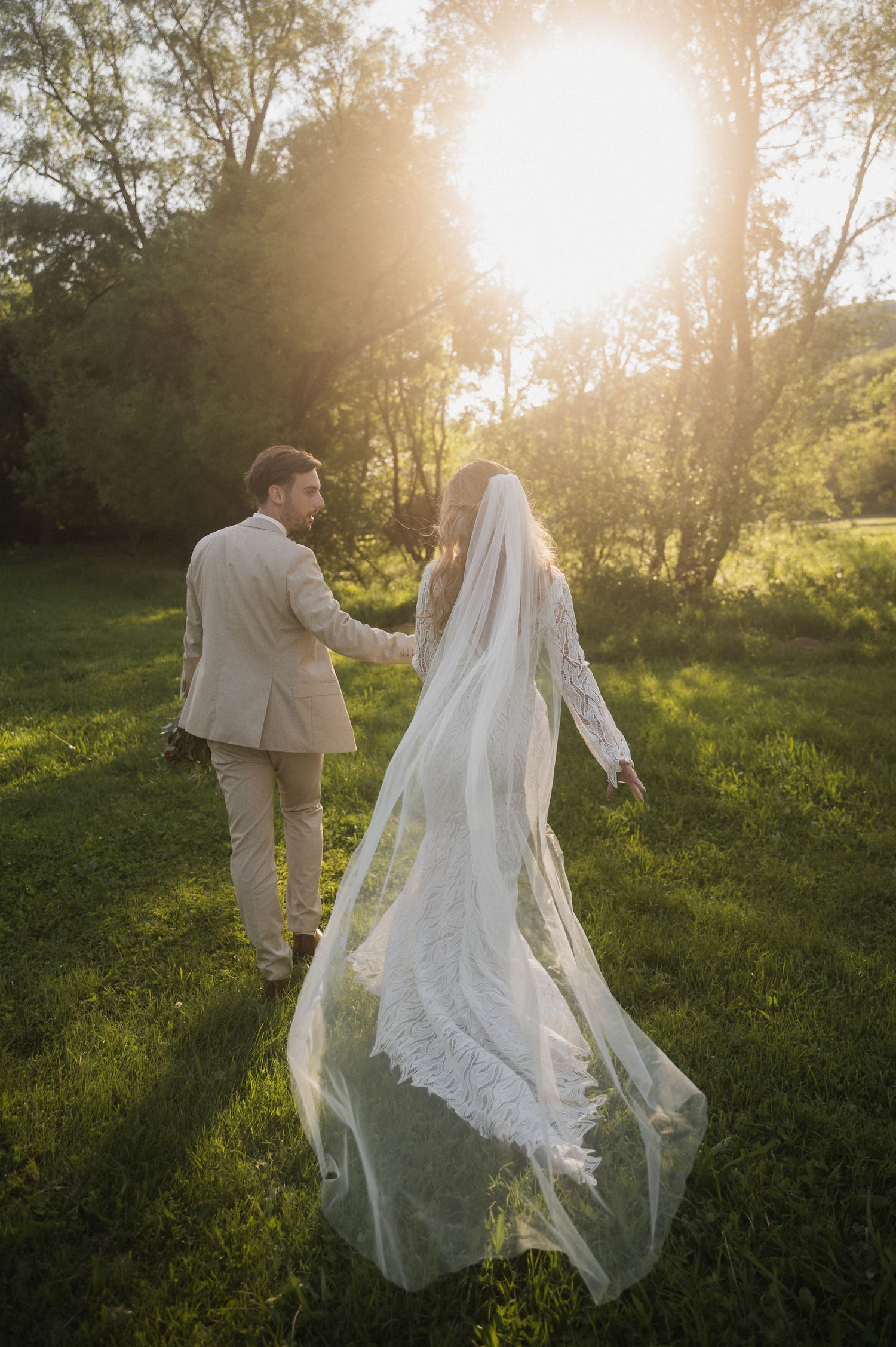 Couple walking through a sunlit meadow at golden hour with flowing white wedding dress and veil catching the breeze.