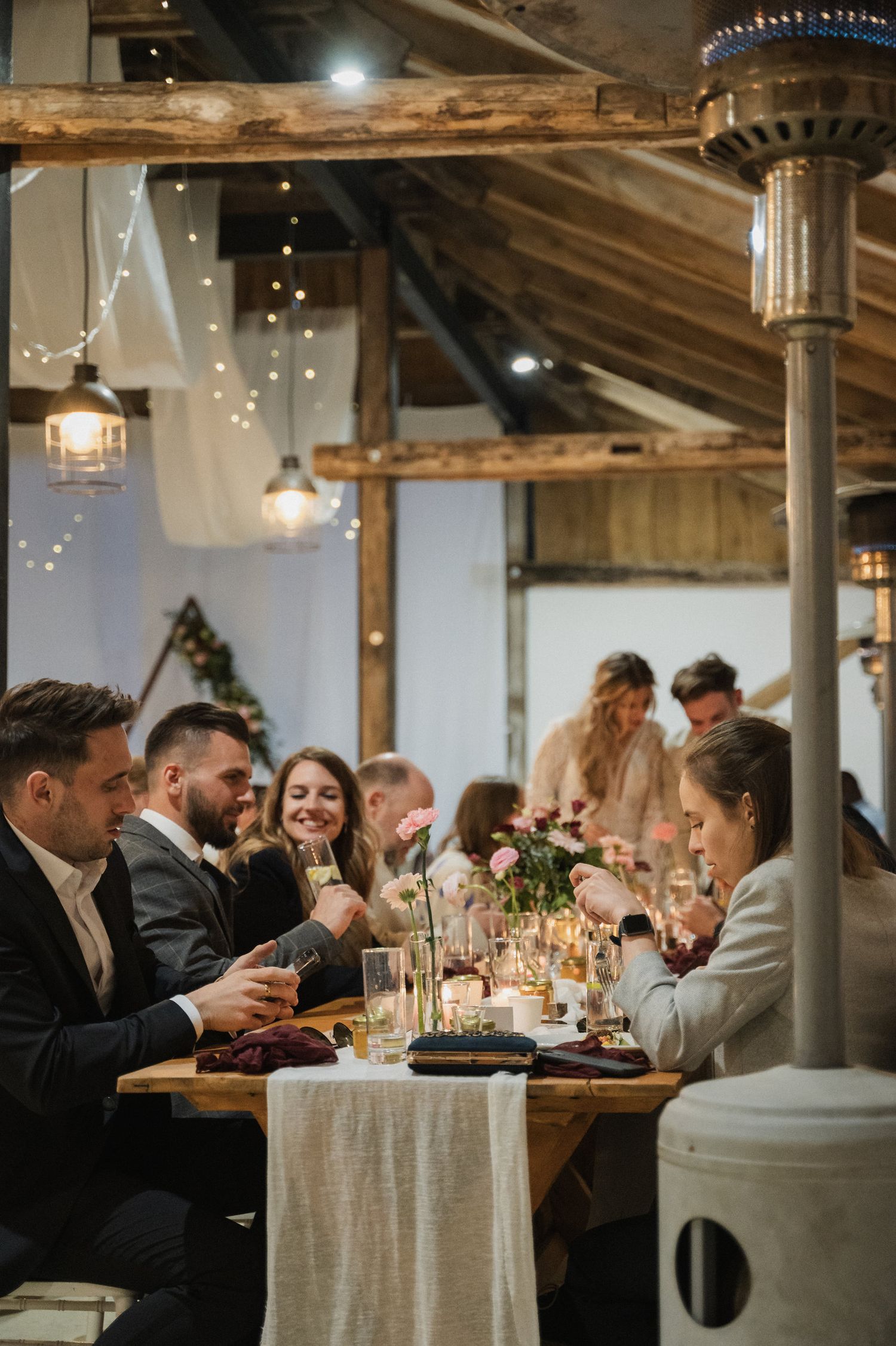 Wedding guests enjoying an elegant dinner reception at a rustic venue with string lights and wooden beams overhead.