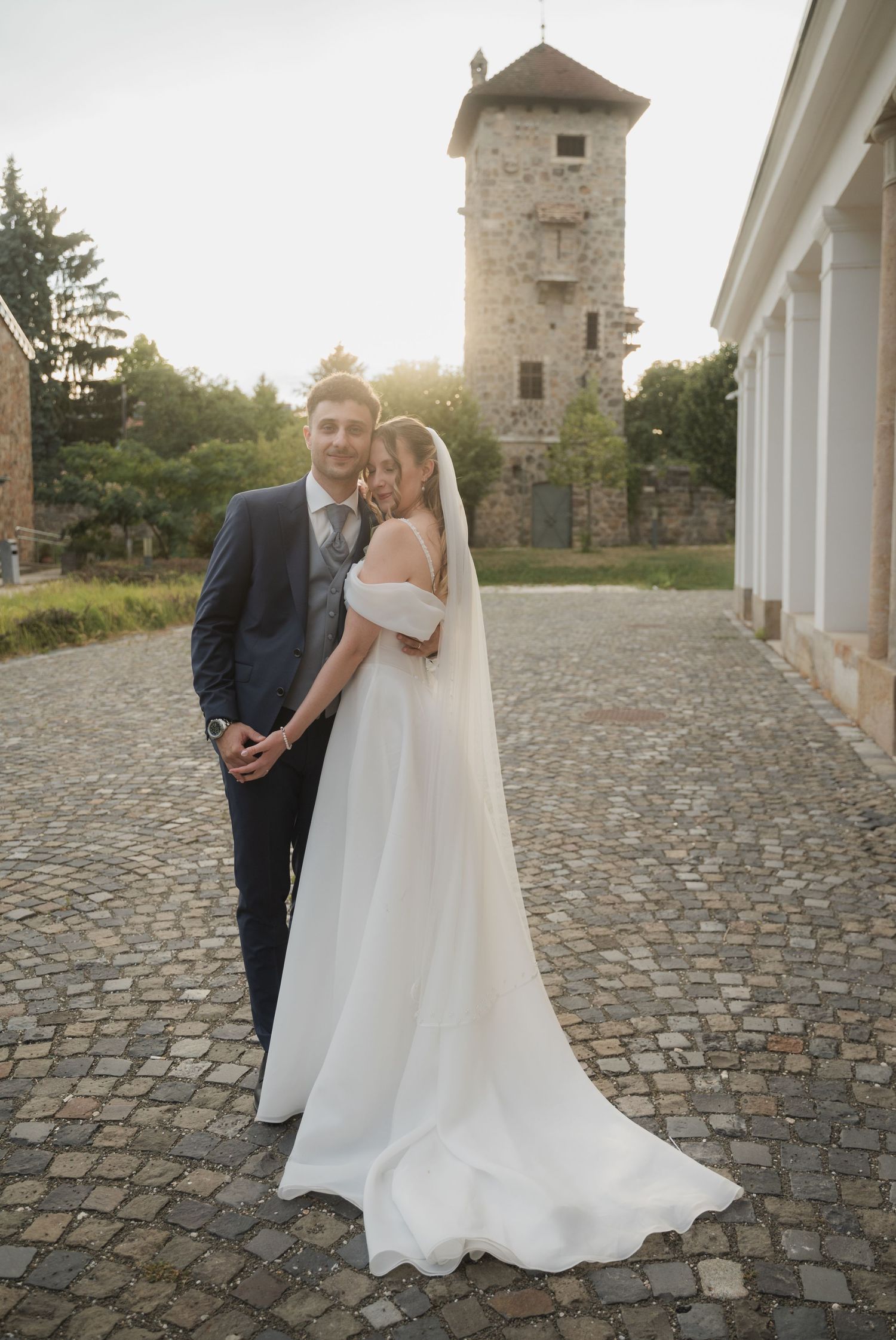 Bride and groom pose on cobblestone courtyard with historic stone tower backdrop at golden hour wedding.