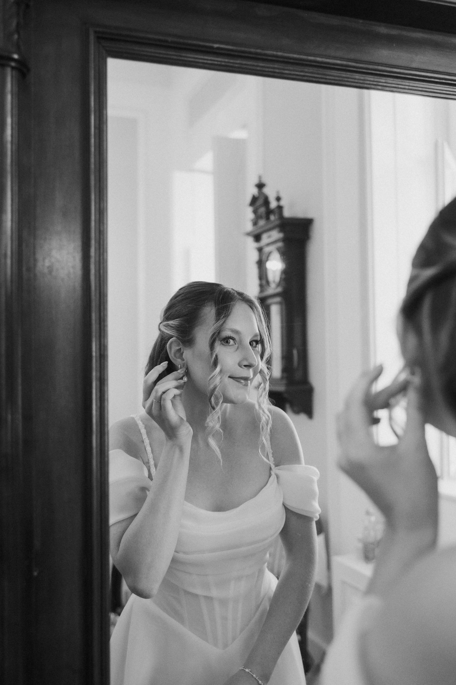 Bride in white gown adjusting earring while gazing into ornate mirror, captured in elegant black and white photography.