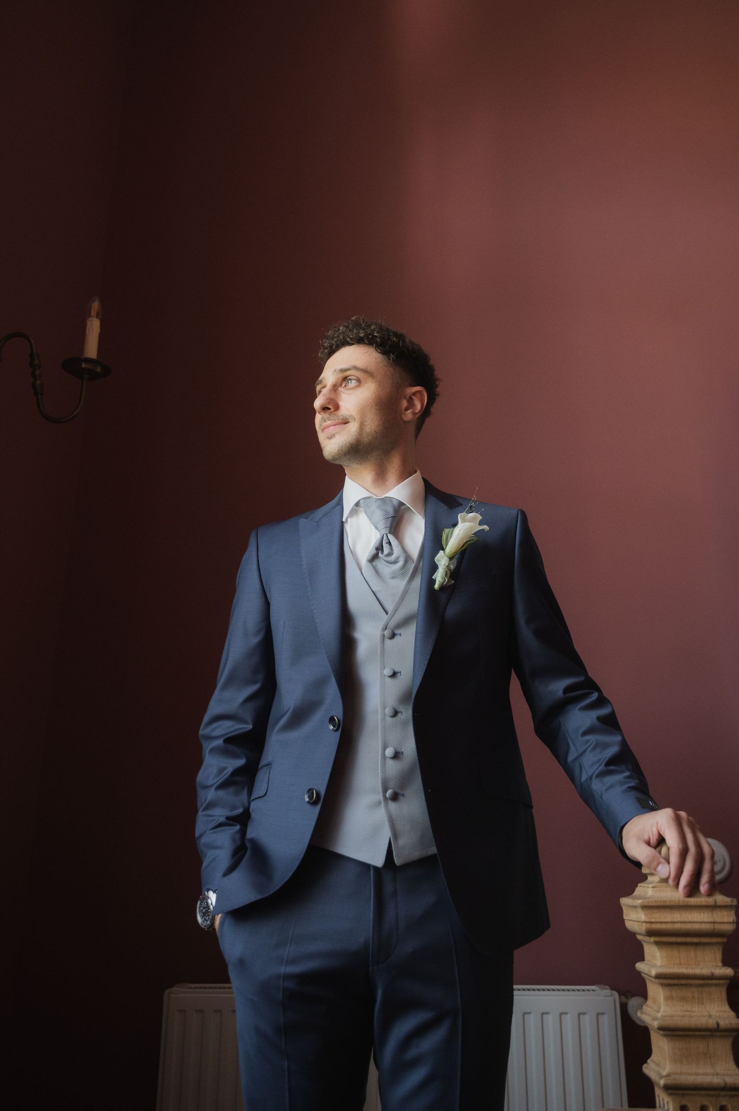 Groom in blue suit with gray vest and boutonniere poses confidently near a fireplace mantel against a deep red wall.