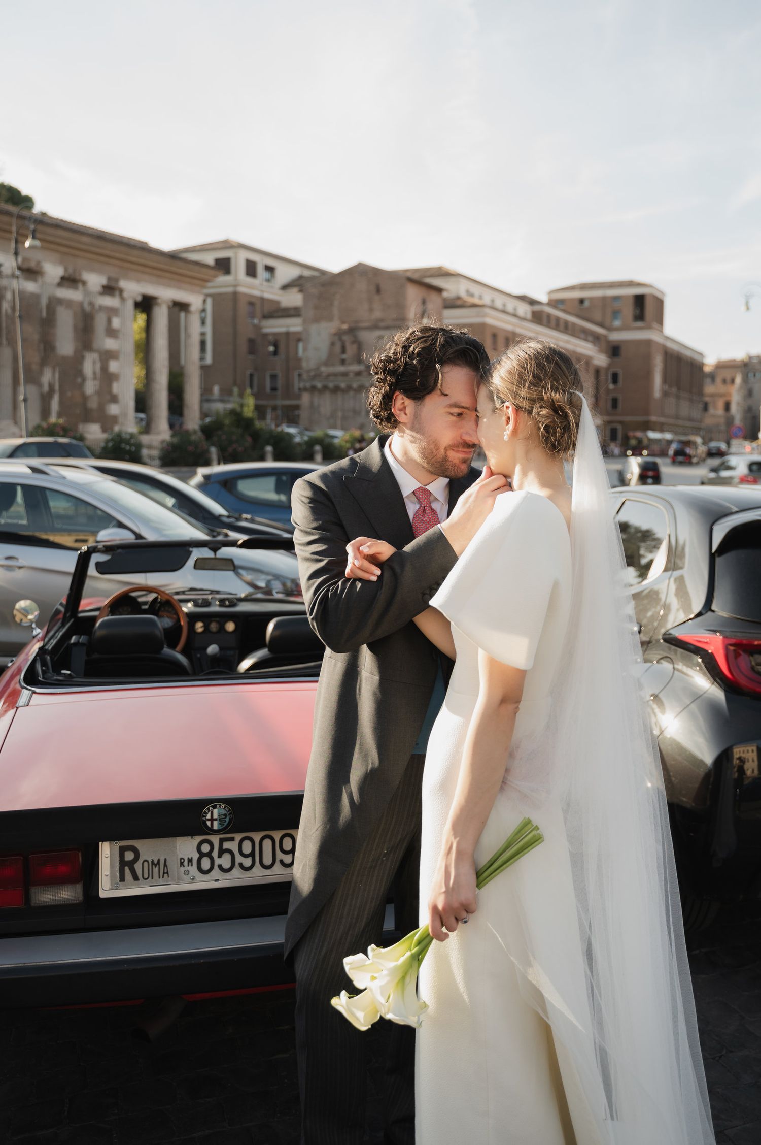 Newlyweds share romantic moment beside classic red convertible car in Italian setting.