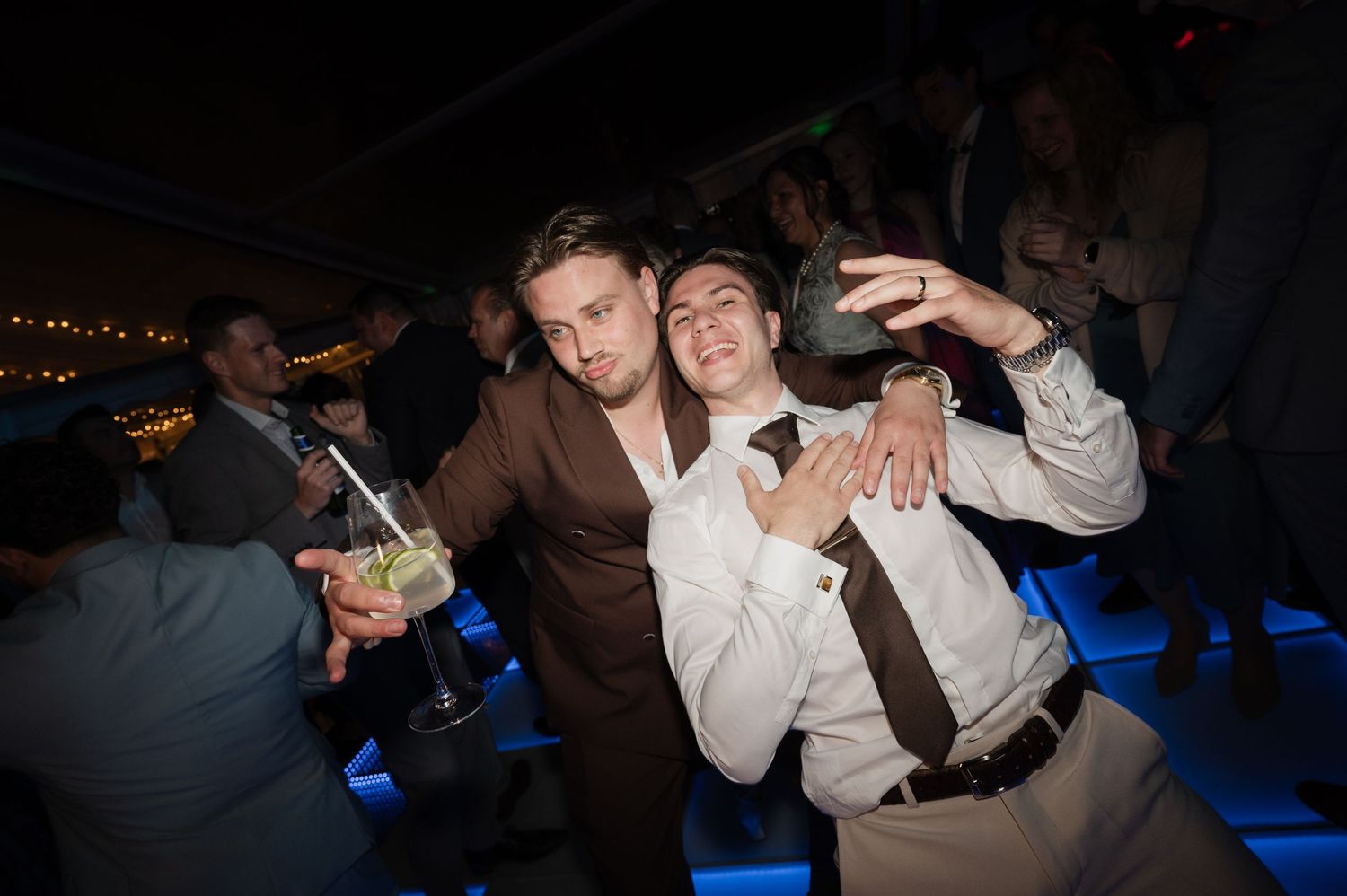 Two young men in suits pose playfully at a lively evening wedding reception dance floor.