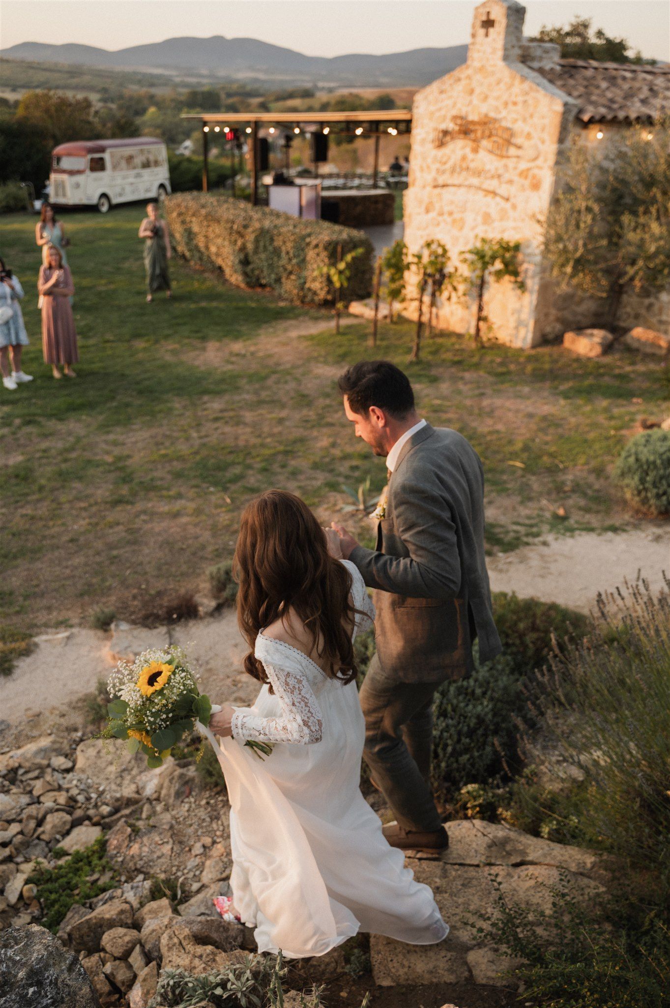 Rustic countryside wedding scene with stone building and string lights at sunset.