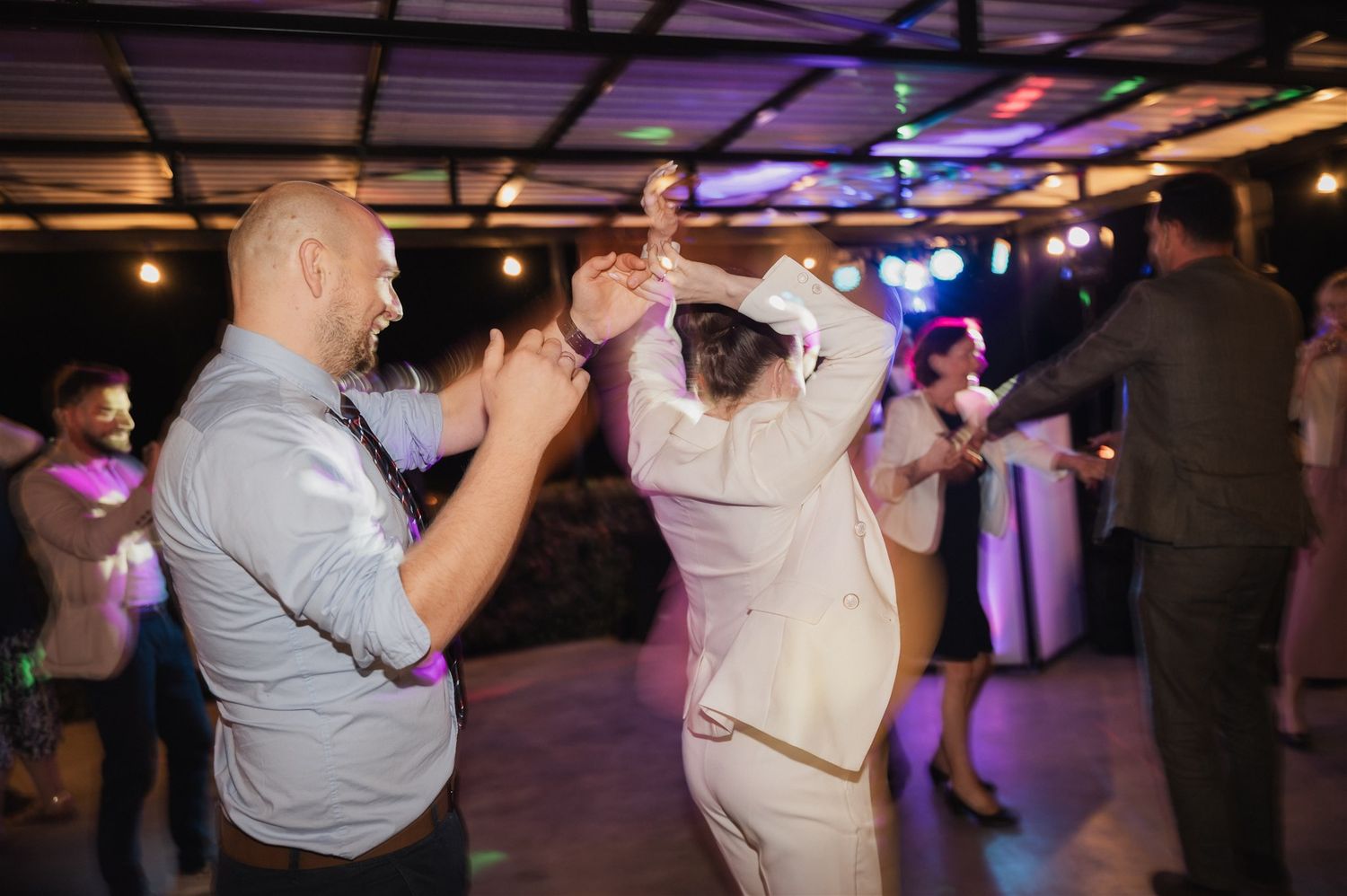 Wedding guests dance and celebrate under purple lighting at an indoor reception venue.