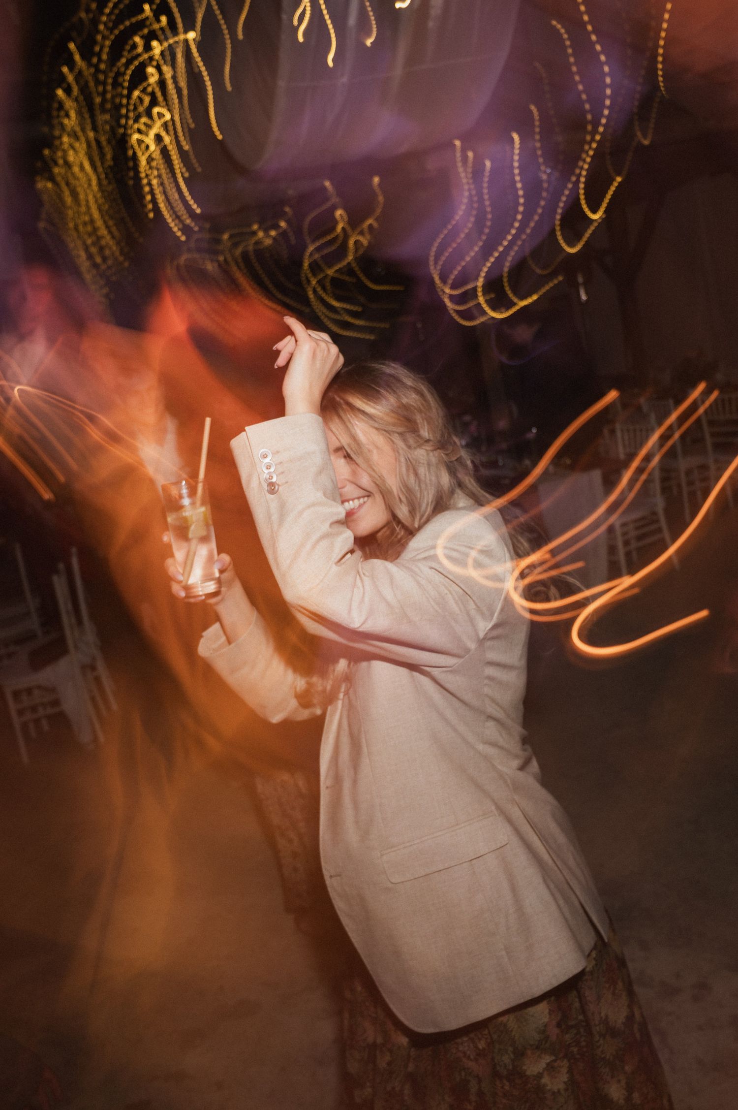 Person in white blazer dancing at a nightclub with swirling light trails and orange glow in the background.