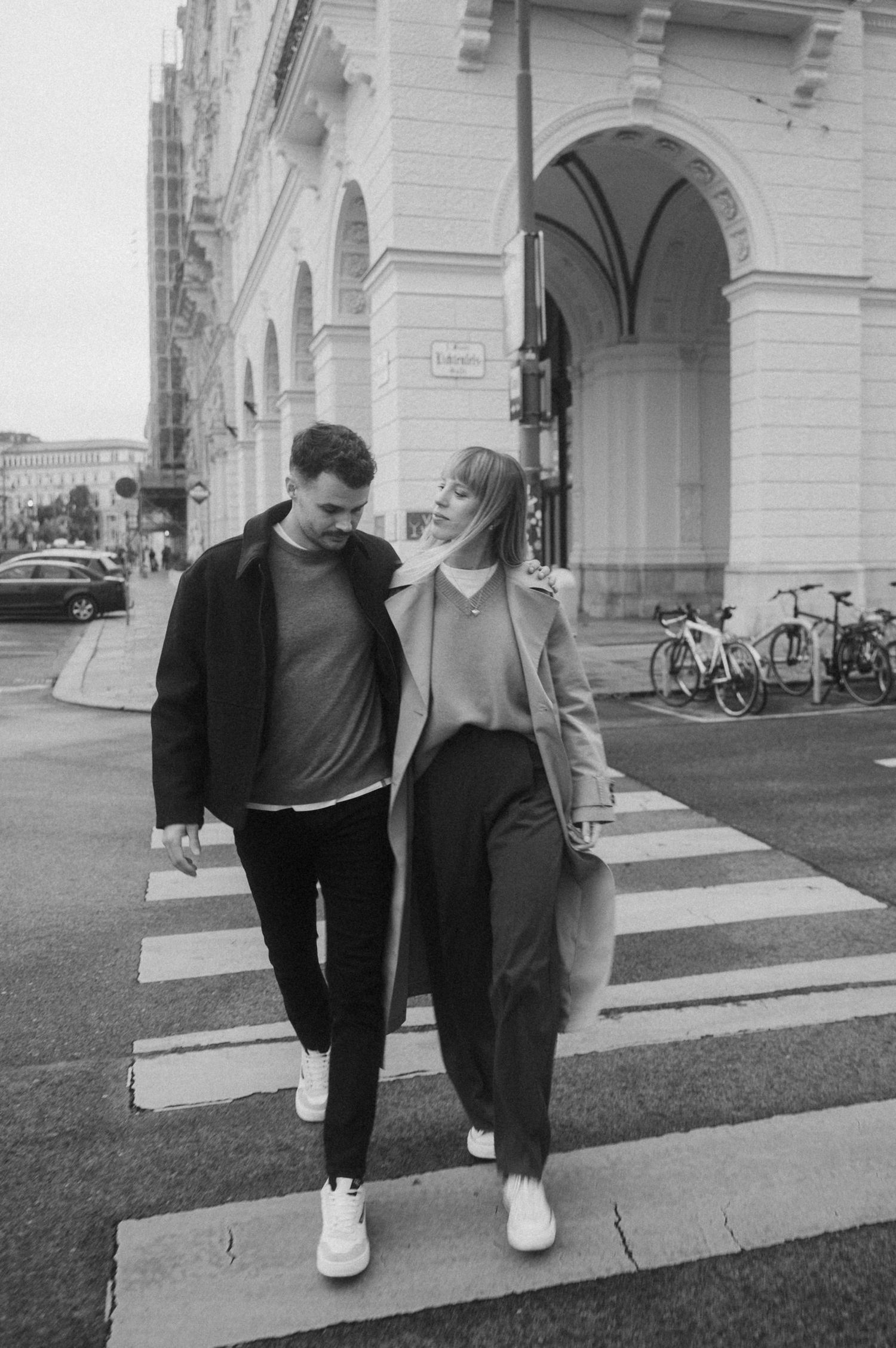 Black and white photo of a couple walking together on a crosswalk in front of historic European architecture.