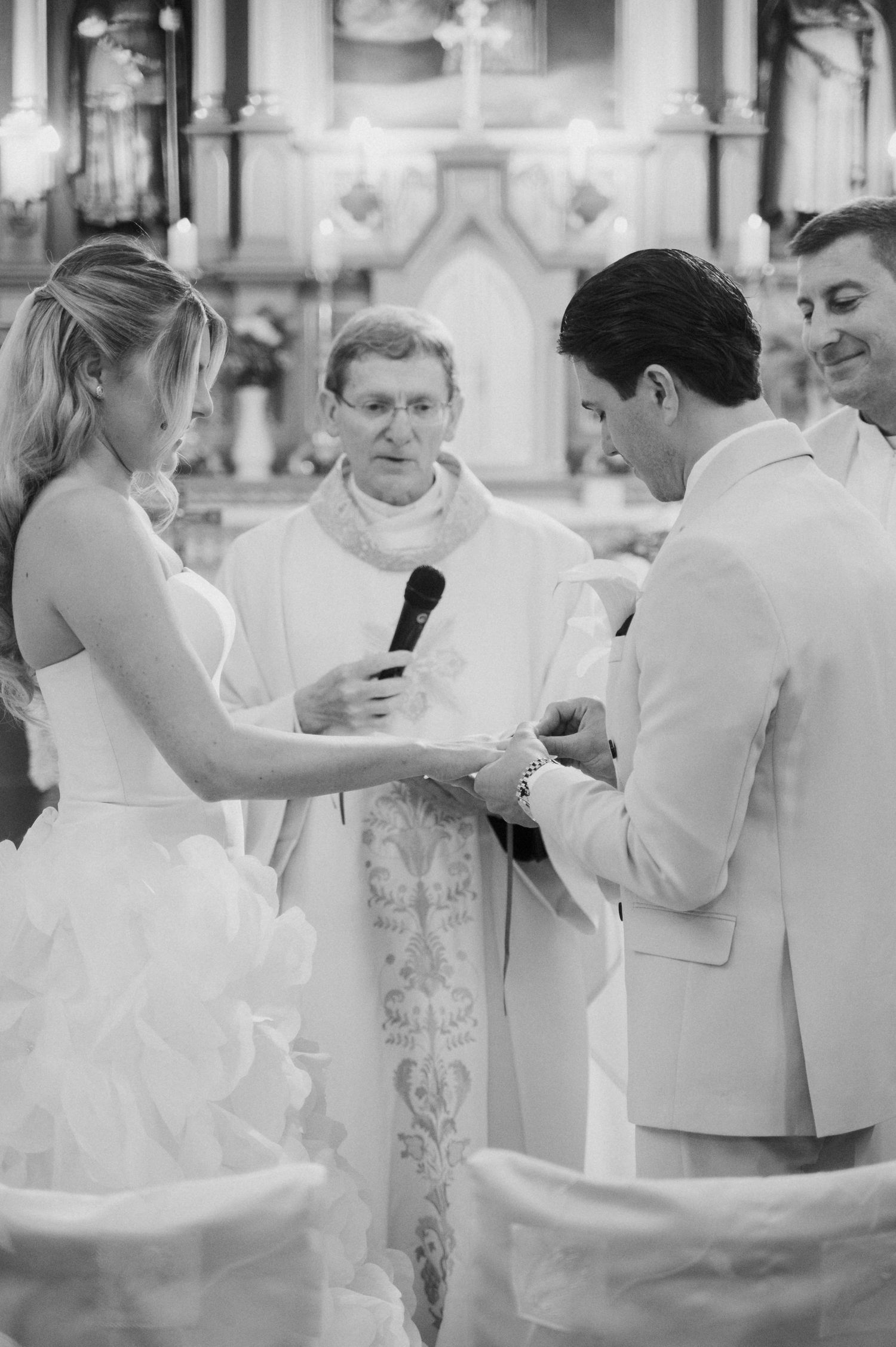 Priest officiating ring exchange ceremony between bride and groom at church altar in black and white.