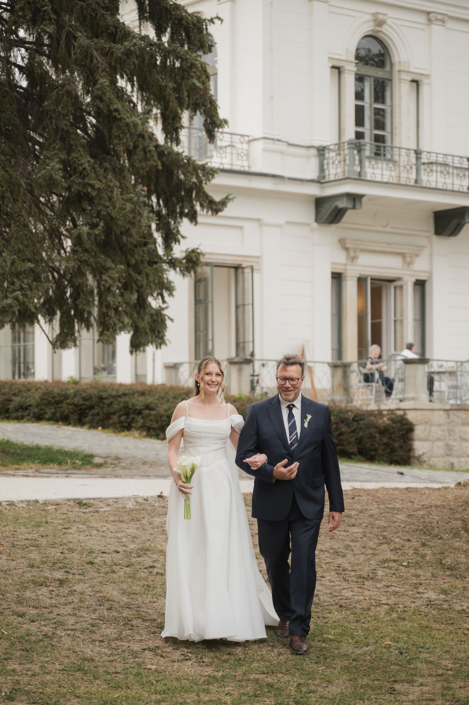 Bride in white gown walks with father toward elegant white mansion on wedding day.