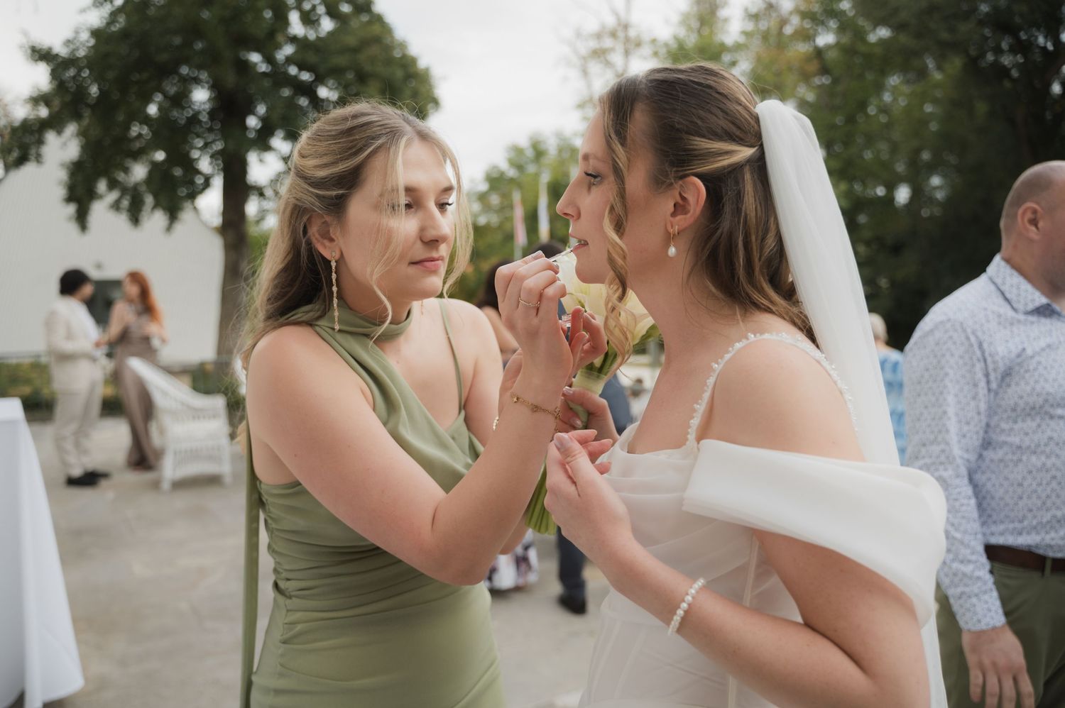 Bridesmaid in green dress helps bride in white gown with boutonniere at outdoor wedding reception.