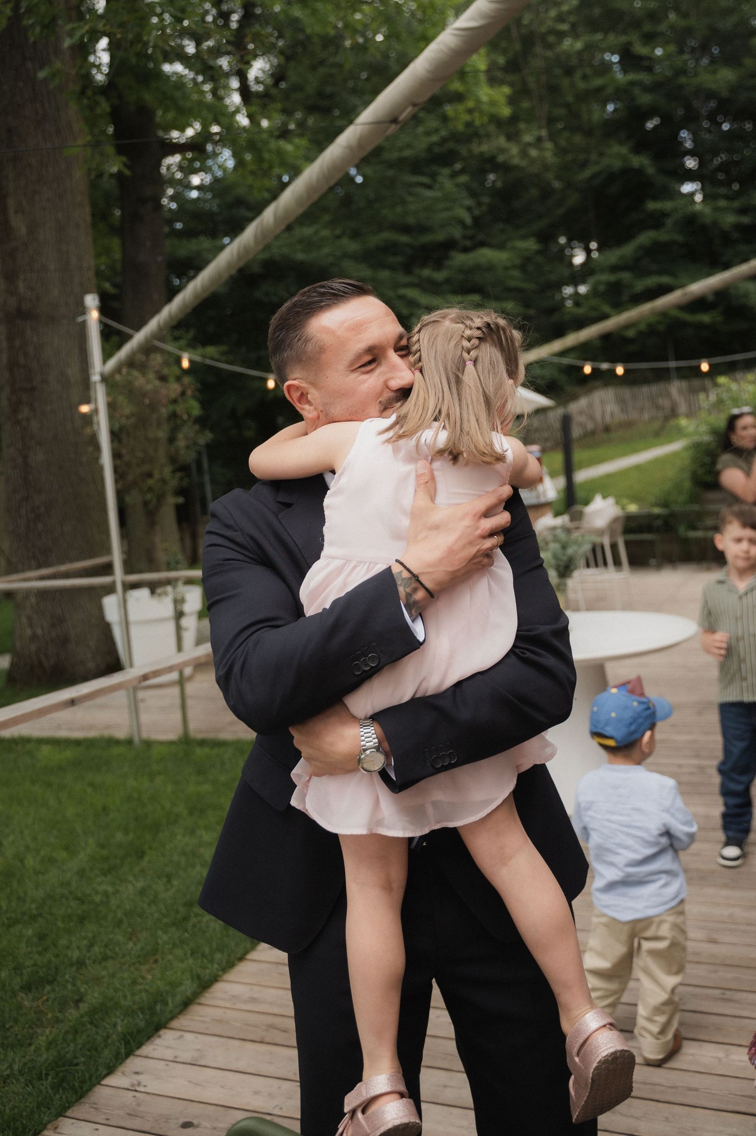 Man in black suit joyfully hugs young girl in pink dress at outdoor wedding reception.