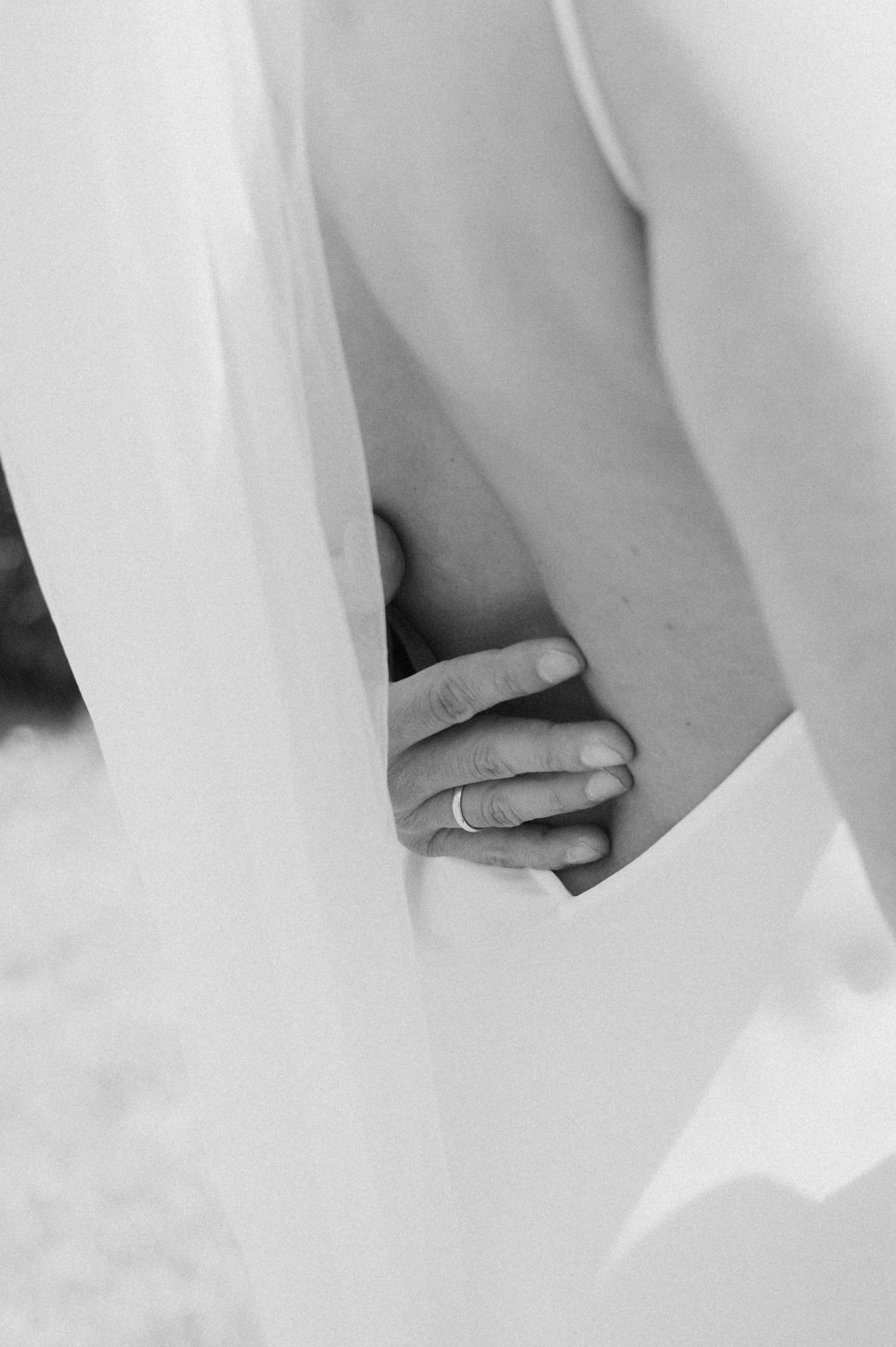 Close-up black and white photo of a groom's hand with wedding ring resting on bride's waist.