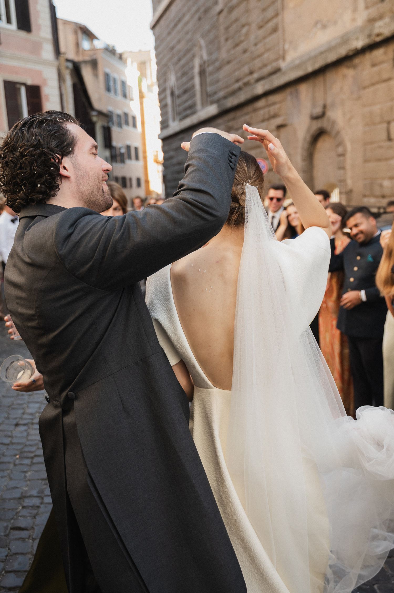 Wedding celebration on cobblestone street with guests throwing confetti in afternoon light.