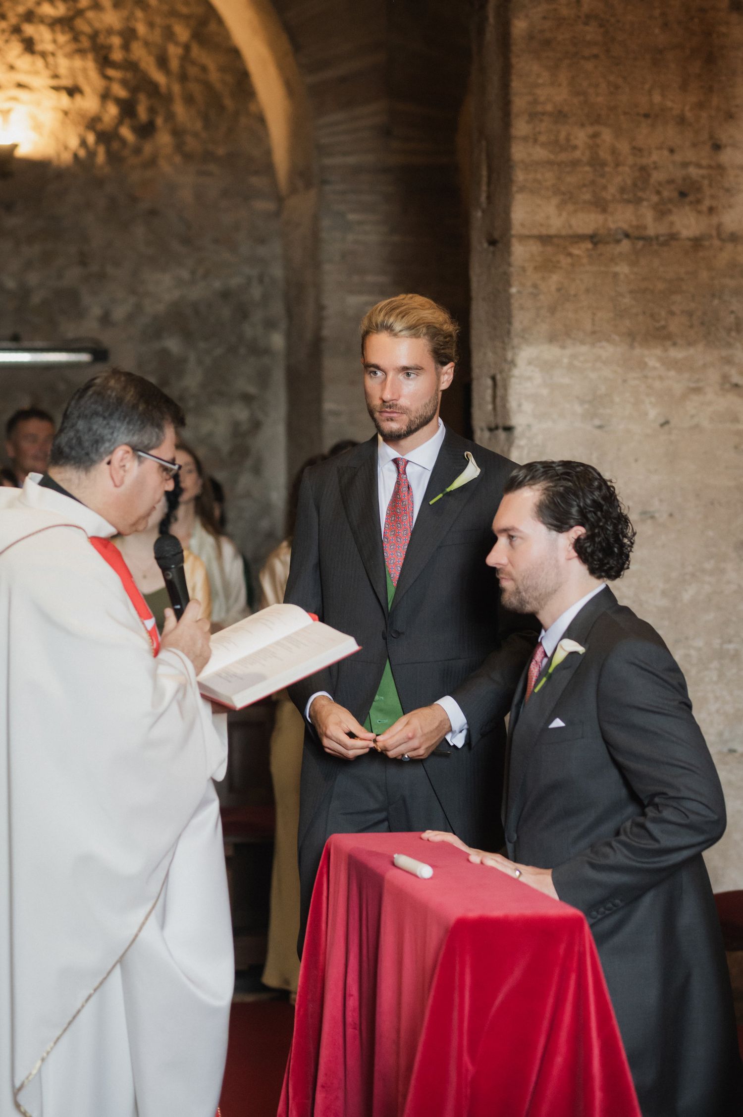 Wedding ceremony participants gather around a red-draped altar in a stone church.