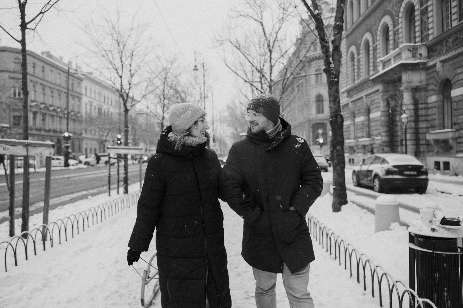 Two people in dark winter coats walking together on a snowy urban street.