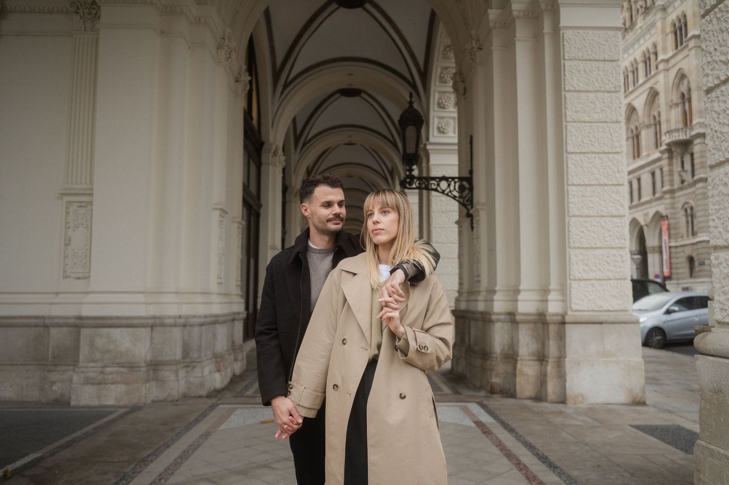 A couple stands together under an ornate archway, with the person in a beige coat holding a small dog.