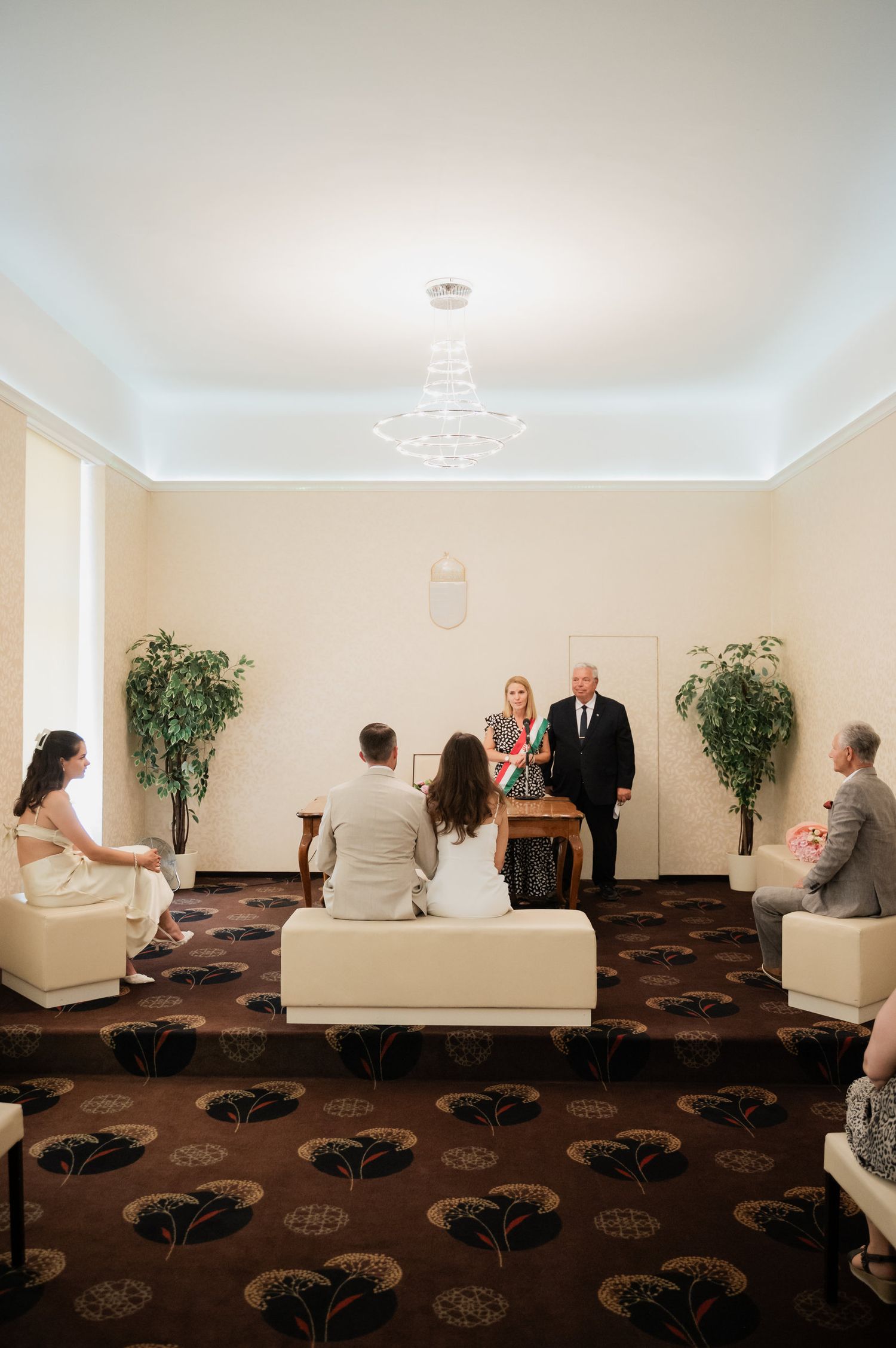 Wedding ceremony setup in a bright white room with brown carpeting and decorative plants.