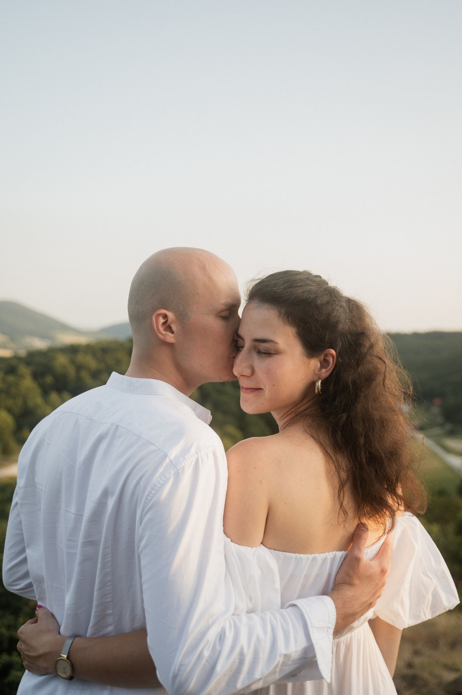 A romantic couple in white clothing embrace while standing on a hillside at sunset.