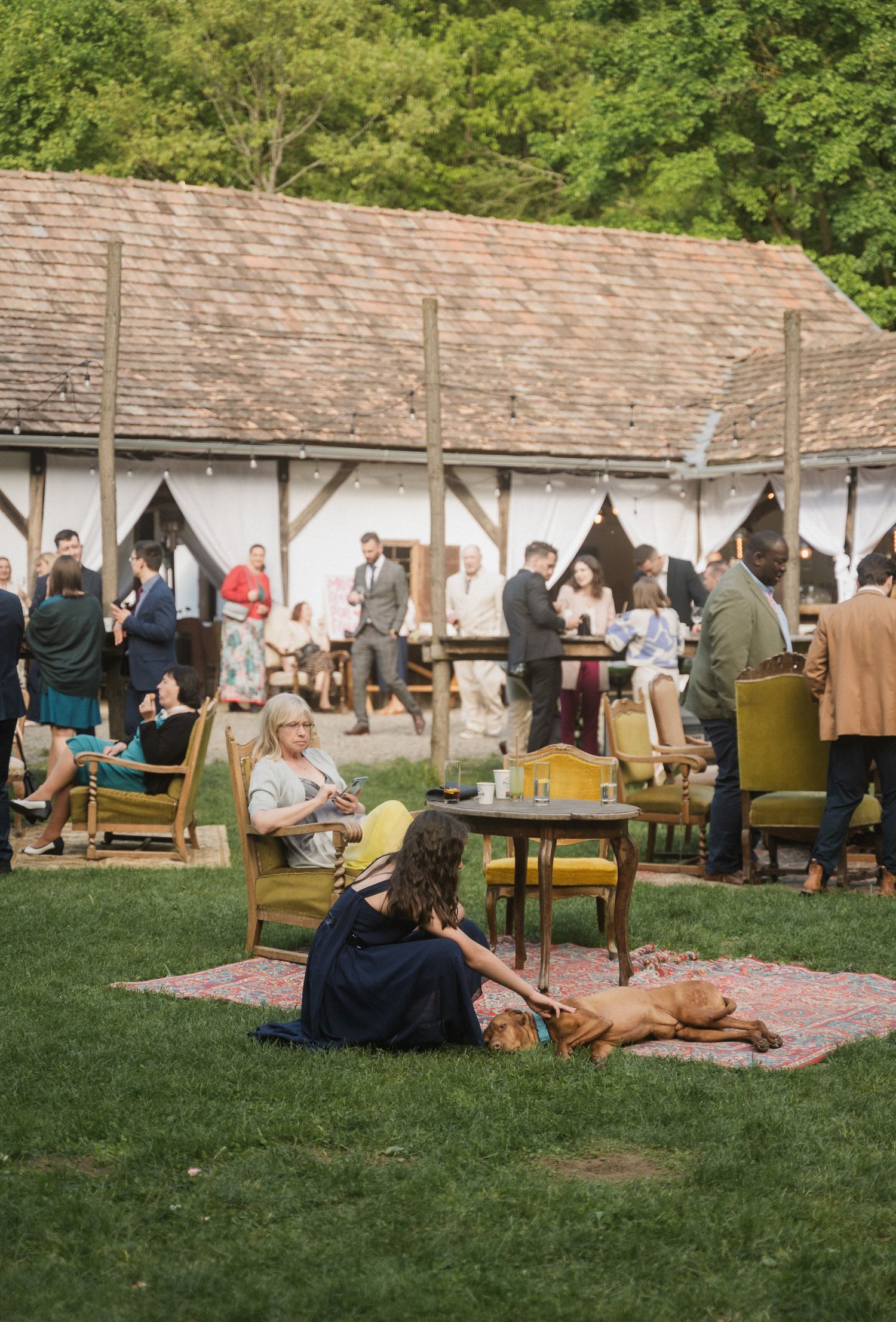 People gather at an outdoor event with vintage furniture and picnic blankets on a grassy lawn next to brick buildings.