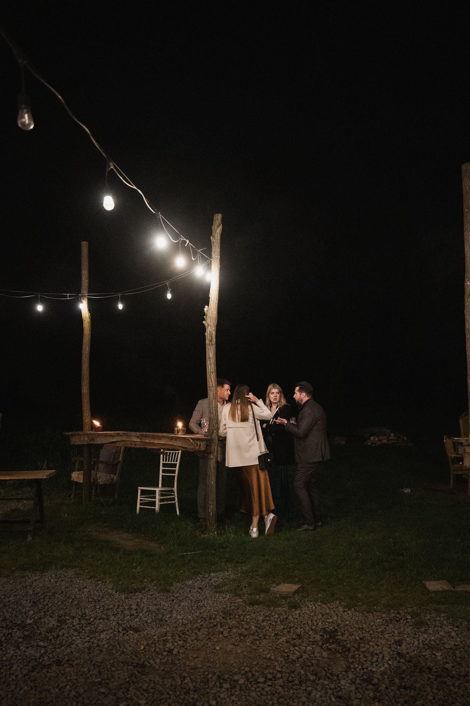 String lights illuminate a dark outdoor gathering area with wooden posts and chairs at night.