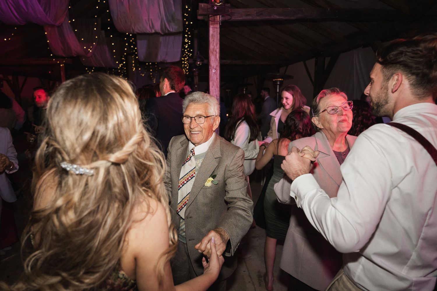 Wedding guests dance and celebrate on a dimly lit dance floor with pink and purple lighting.