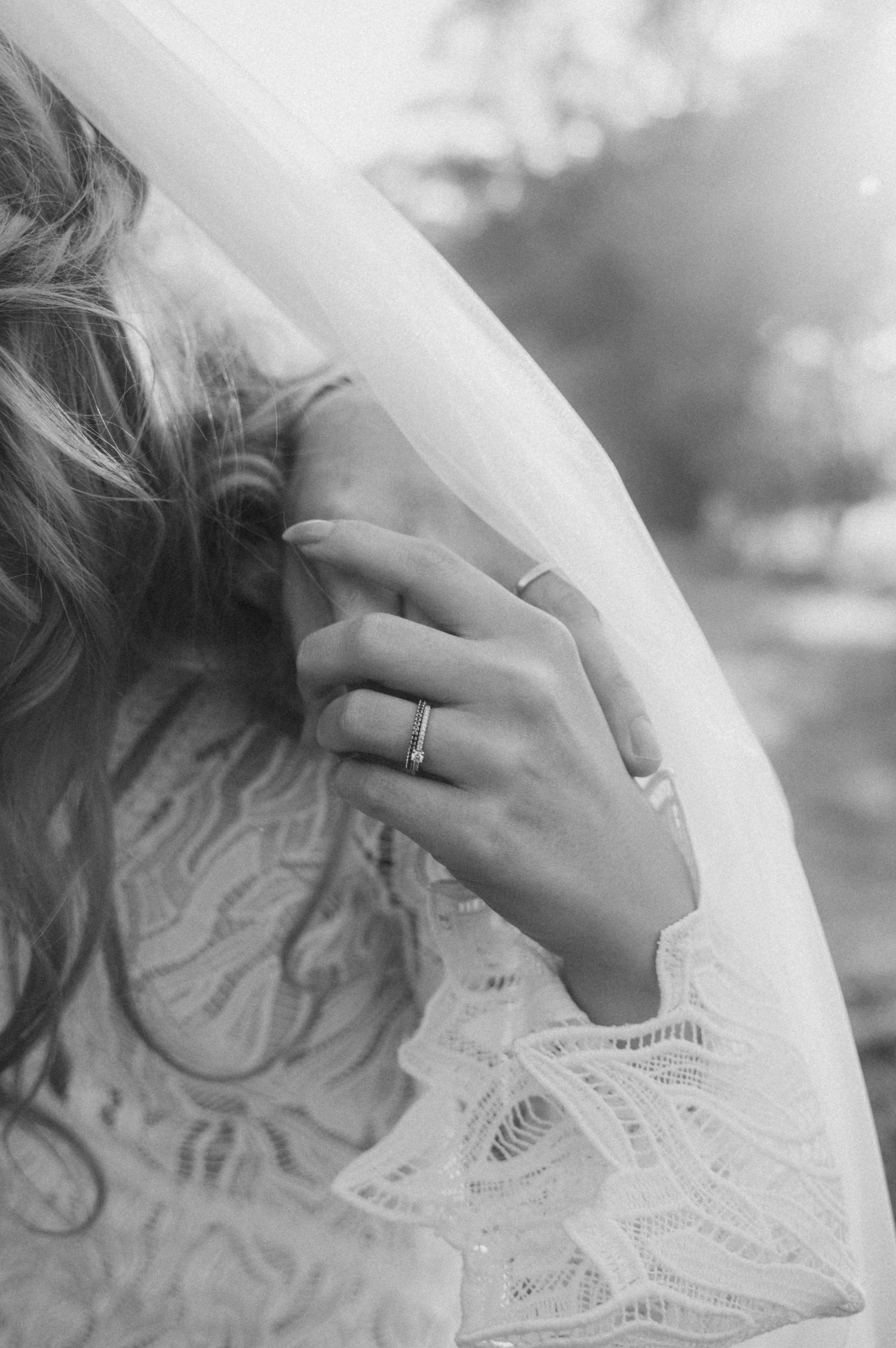 Close-up black and white photograph showing delicate white lace wedding dress details and a hand with a ring.