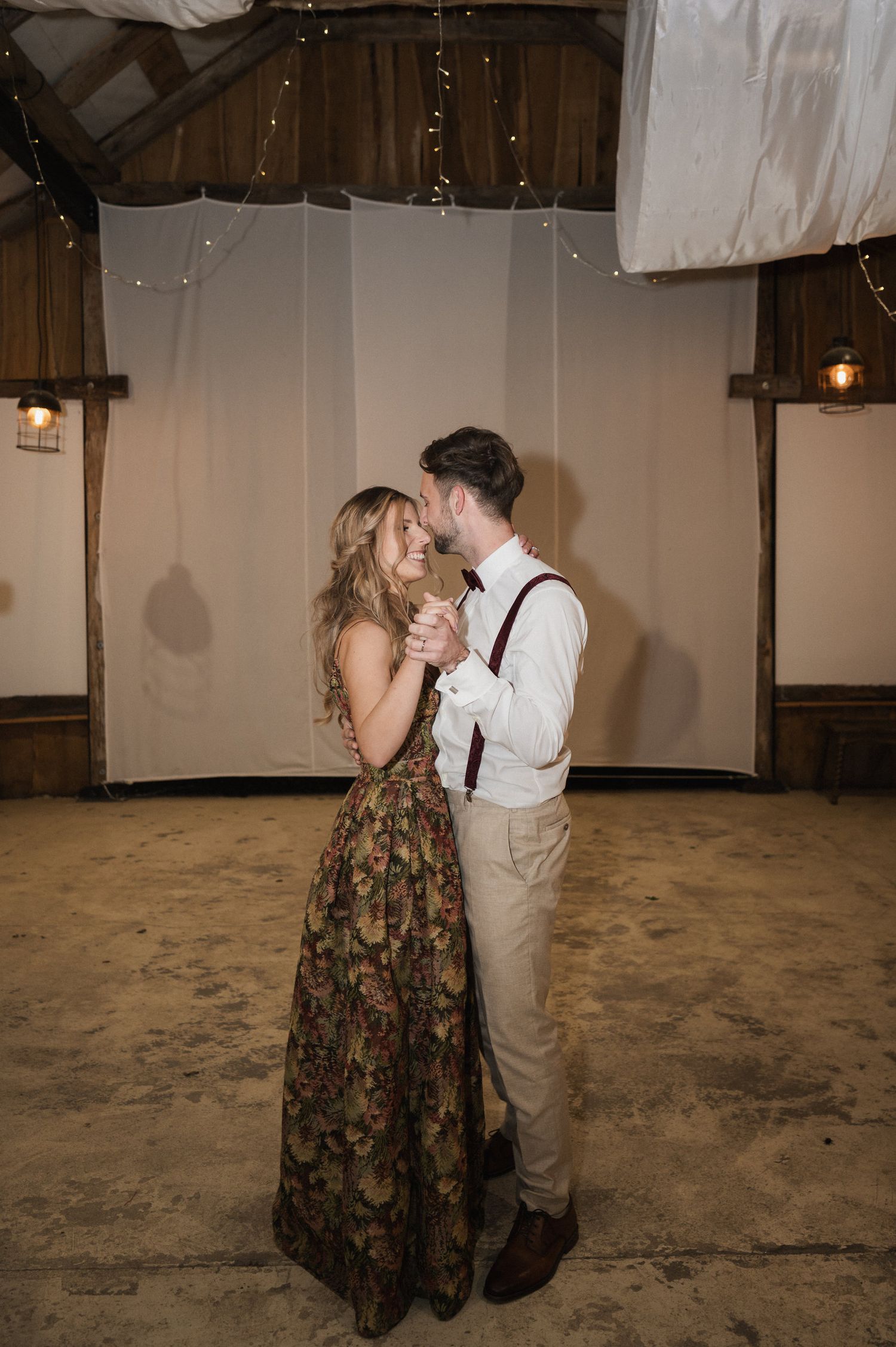 A couple shares a romantic dance moment in a rustic indoor venue with white drapes and exposed beams.