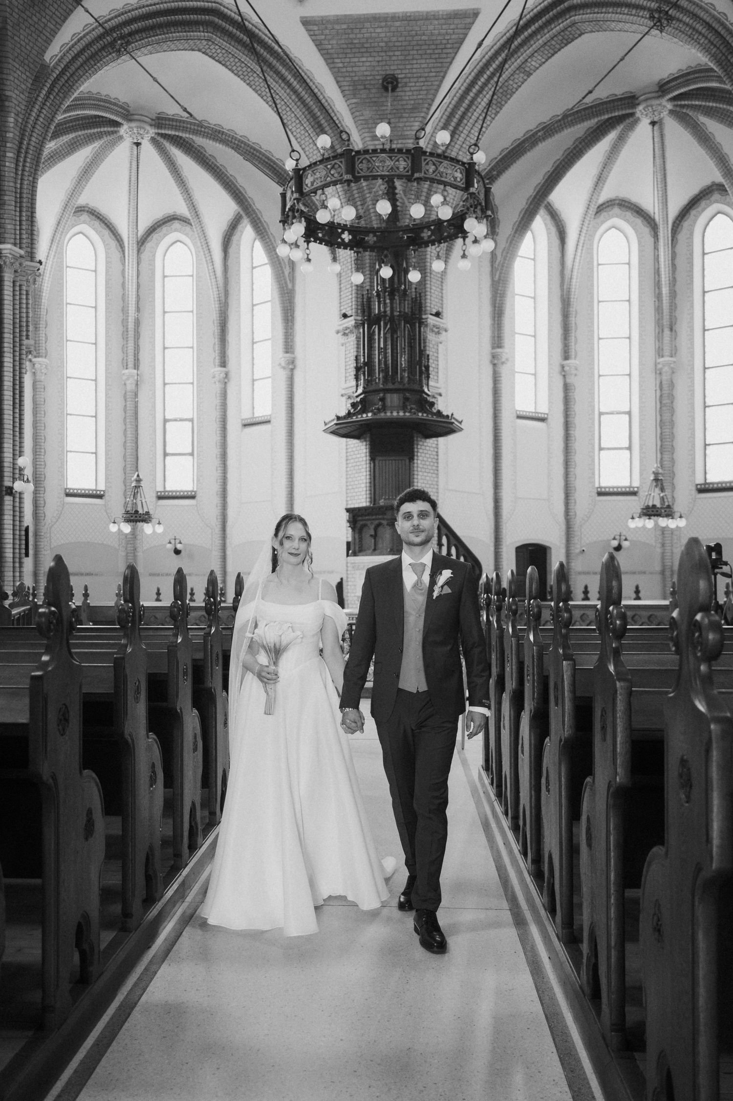 Bride and groom walk down the aisle of an elegant church with arched ceilings in a black and white wedding photo.