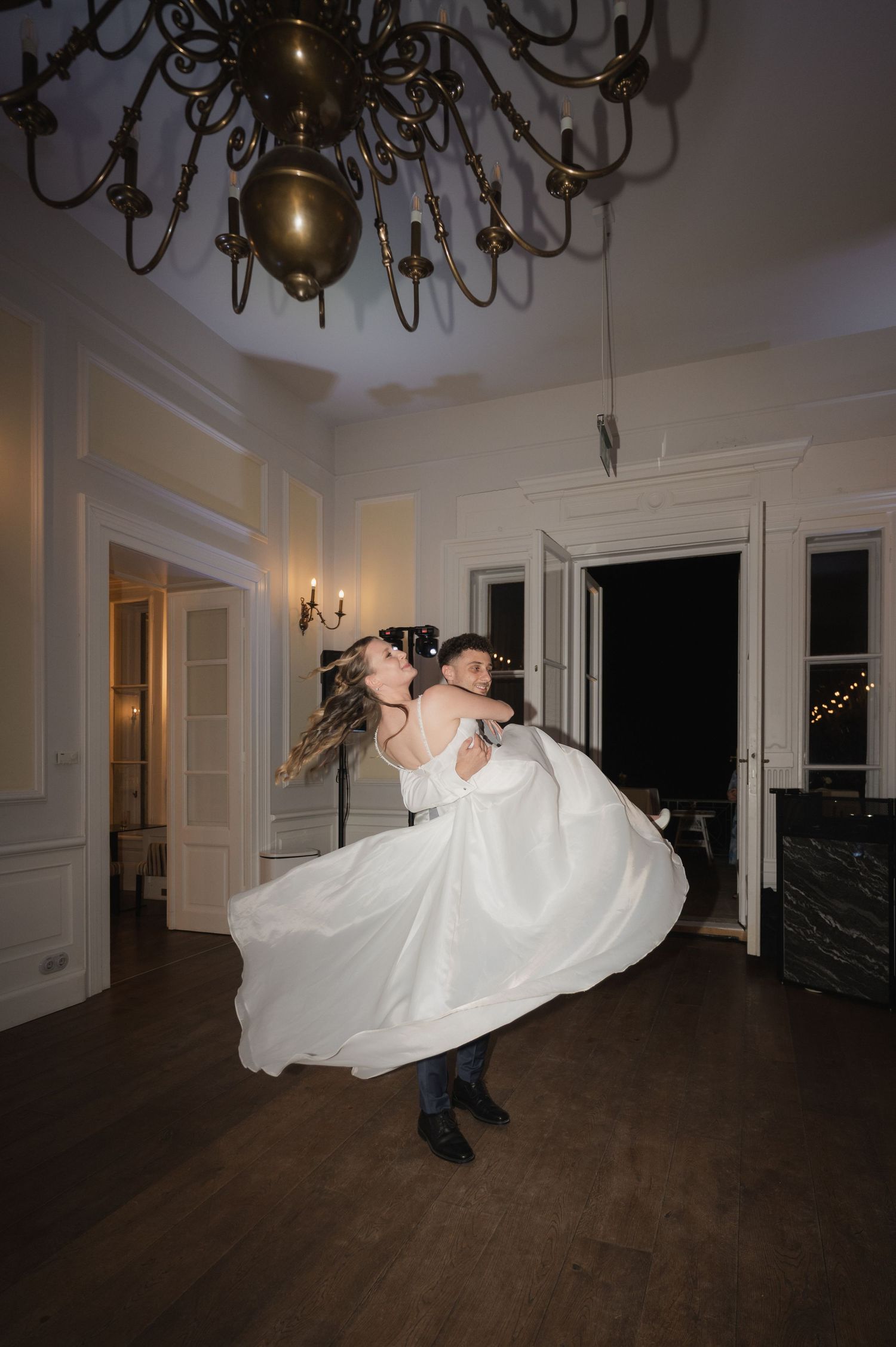 Groom lifting bride in white ball gown during first dance under ornate chandelier in elegant ballroom.
