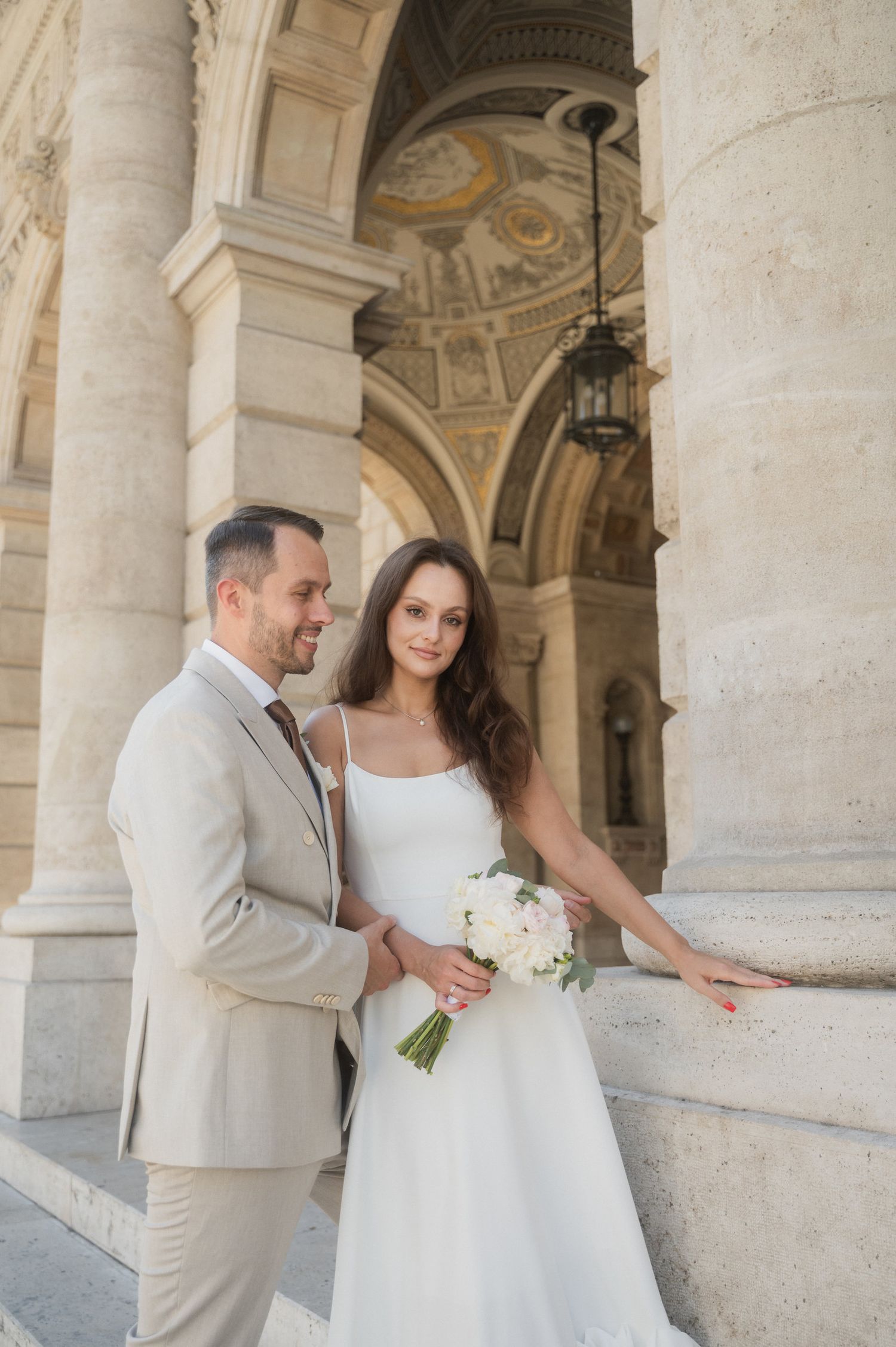 Elegant couple poses beneath classical stone colonnade with ornate ceiling frescoes.