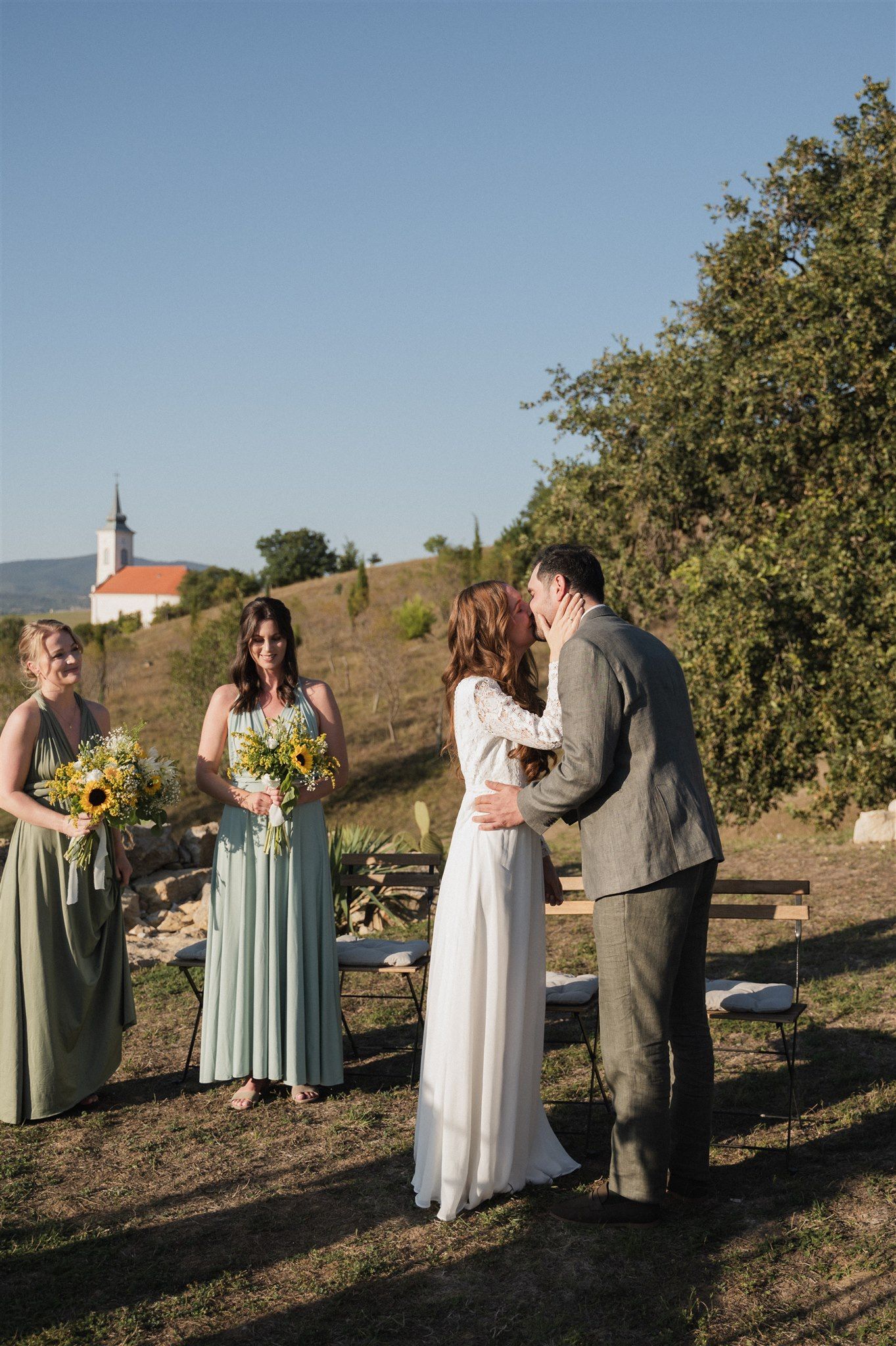 Wedding party poses on hillside with church steeple visible in background.