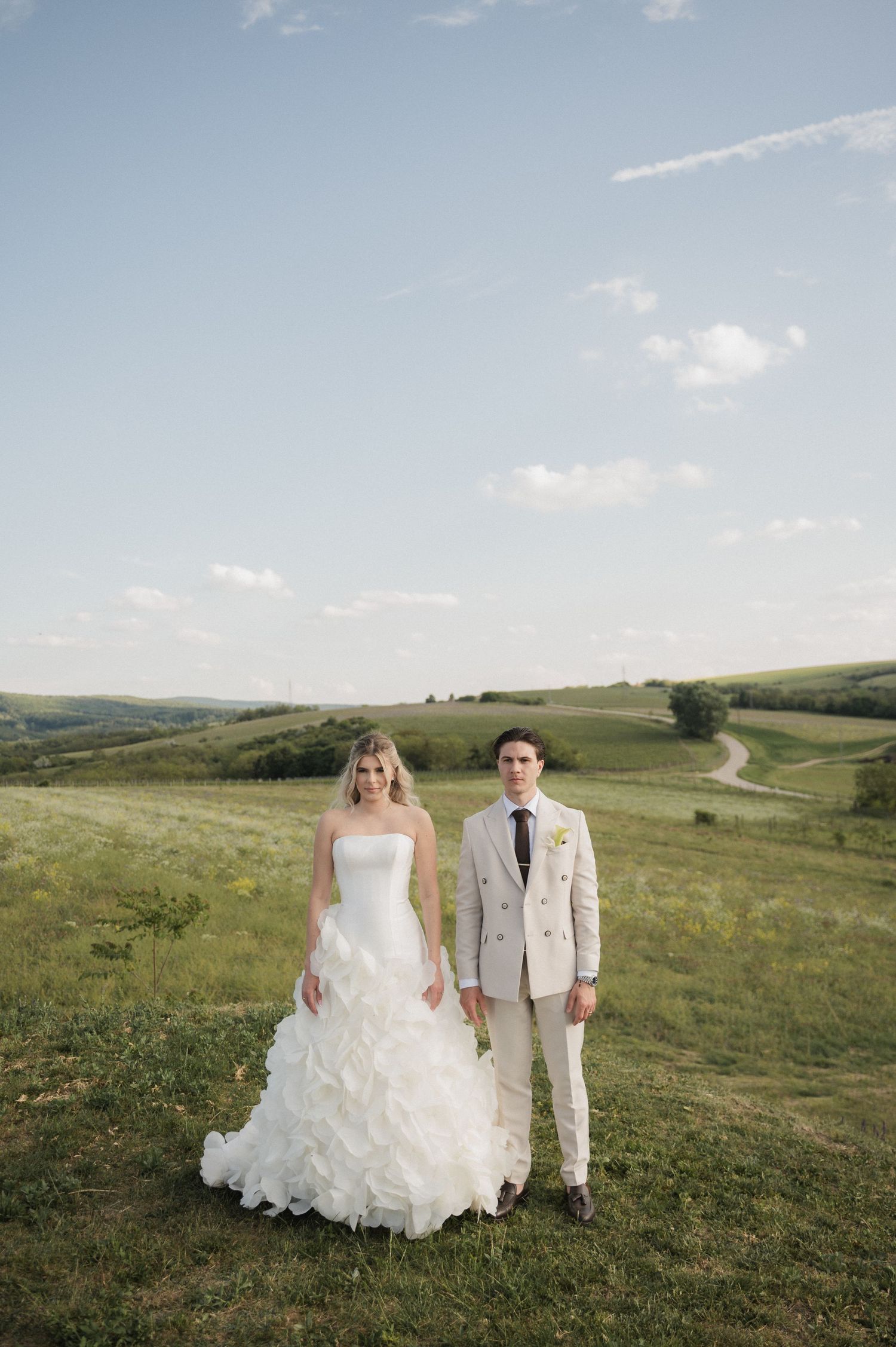 Bride in ruffled strapless gown and groom in beige suit posing in a scenic green countryside.