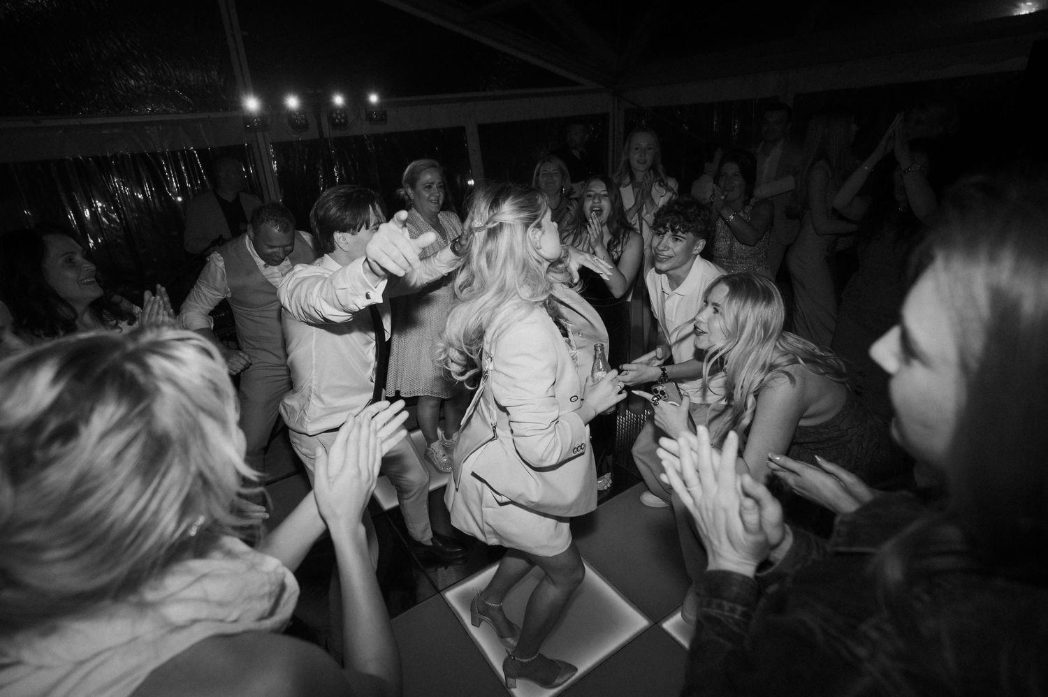 Energetic wedding guests surround bride on crowded dance floor during festive nighttime reception celebration.