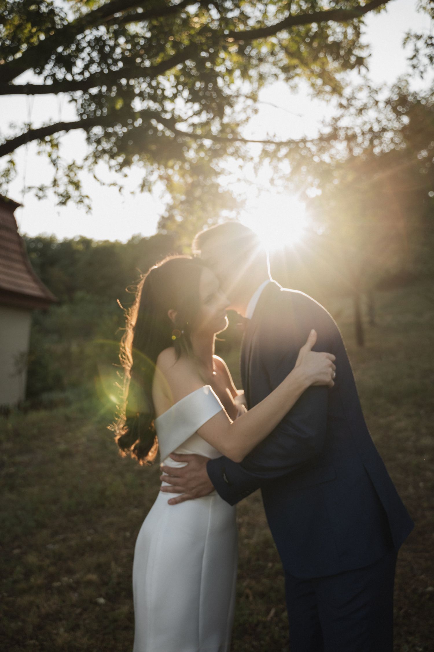 A romantic sunset wedding photo captures a couple embracing outdoors with golden sunlight streaming through trees.
